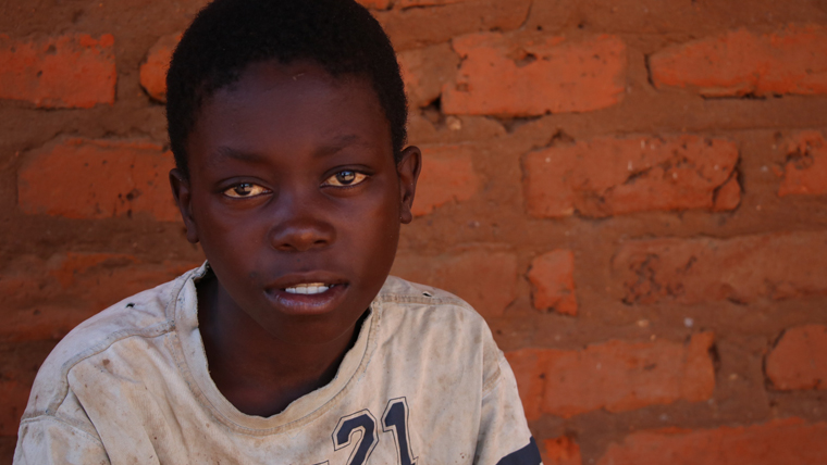 Zambian boy sits in front of a brick wall and looks towards the camera