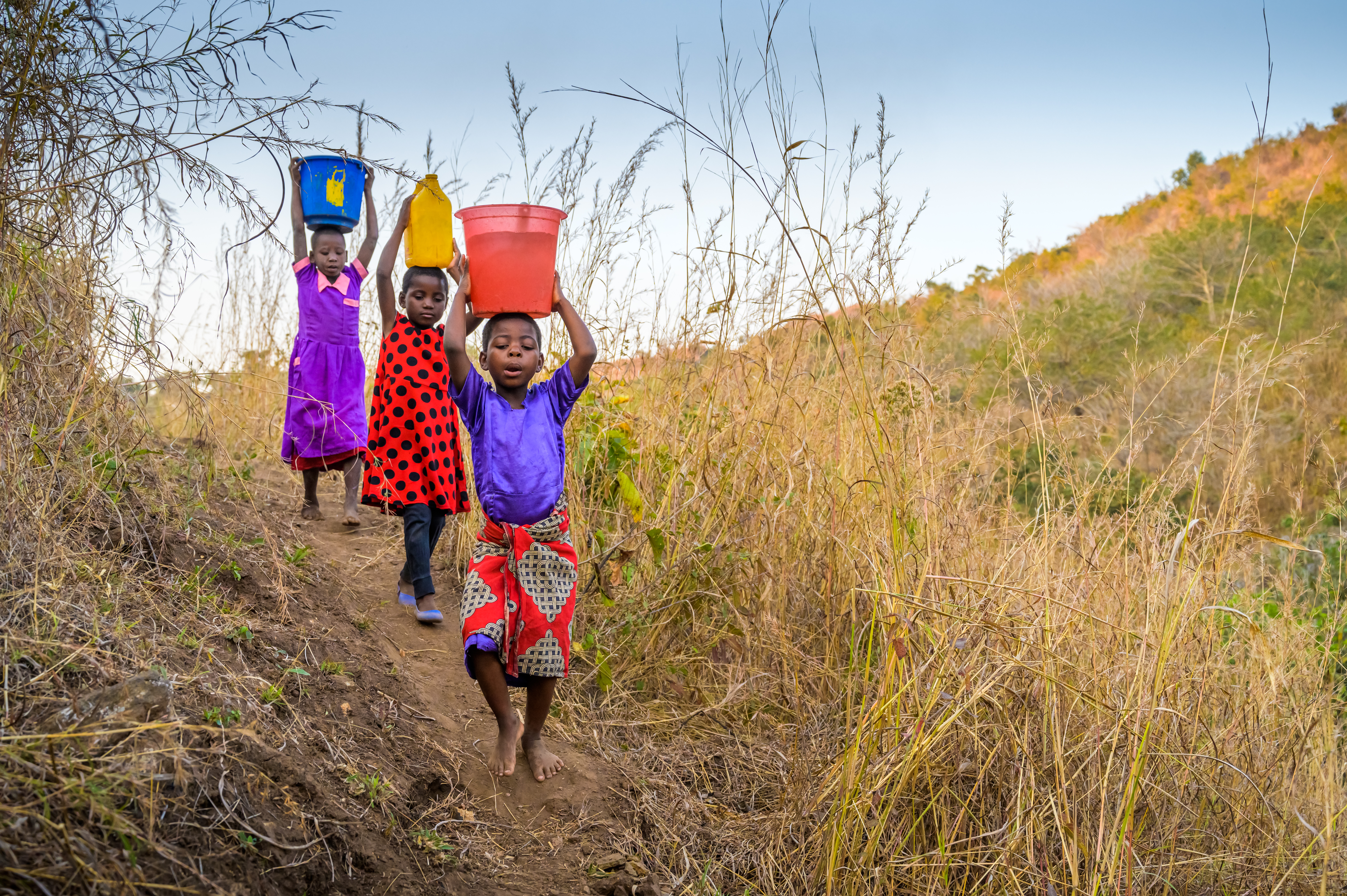 Three children in Malawi walk with water buckets on their head, collecting it from a nearby river
