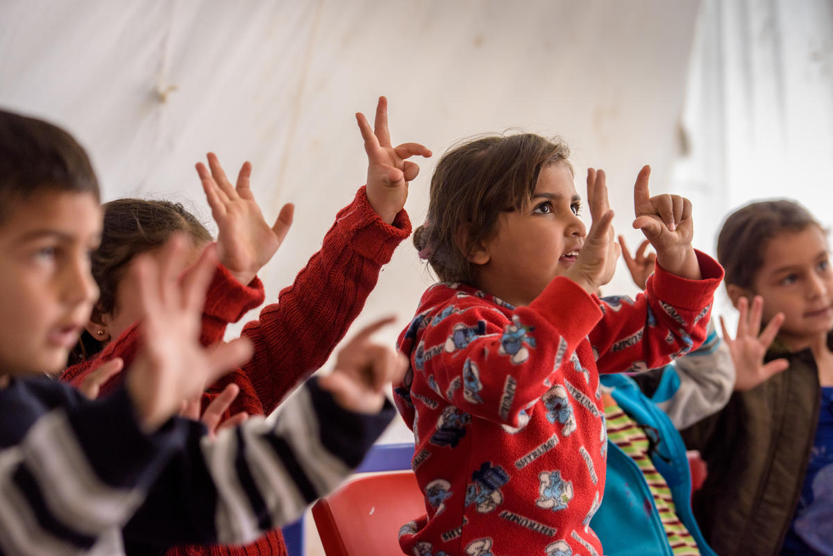Children in a class raising their hands and fingers counting as they look to their teacher (not in view)
