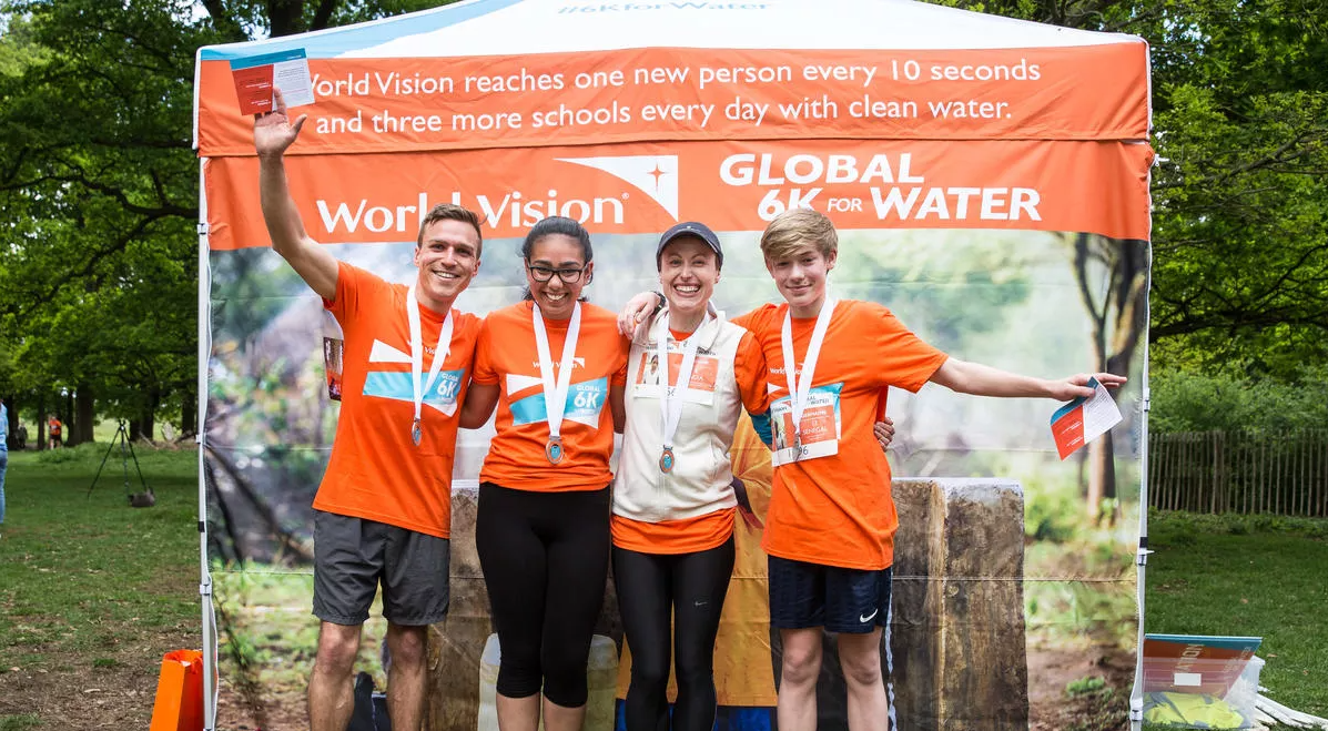 Four people in the UK smile and wave wearing their global 6K medals, standing in front of a pop up tent