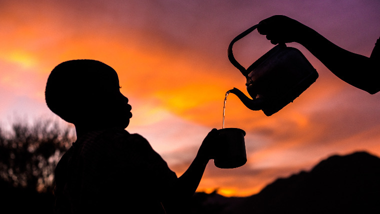 Silhouette of a child in Kenya holding up a mug, with an arm tilting a kettle to pour water into the mug, against the backdrop of a sunset