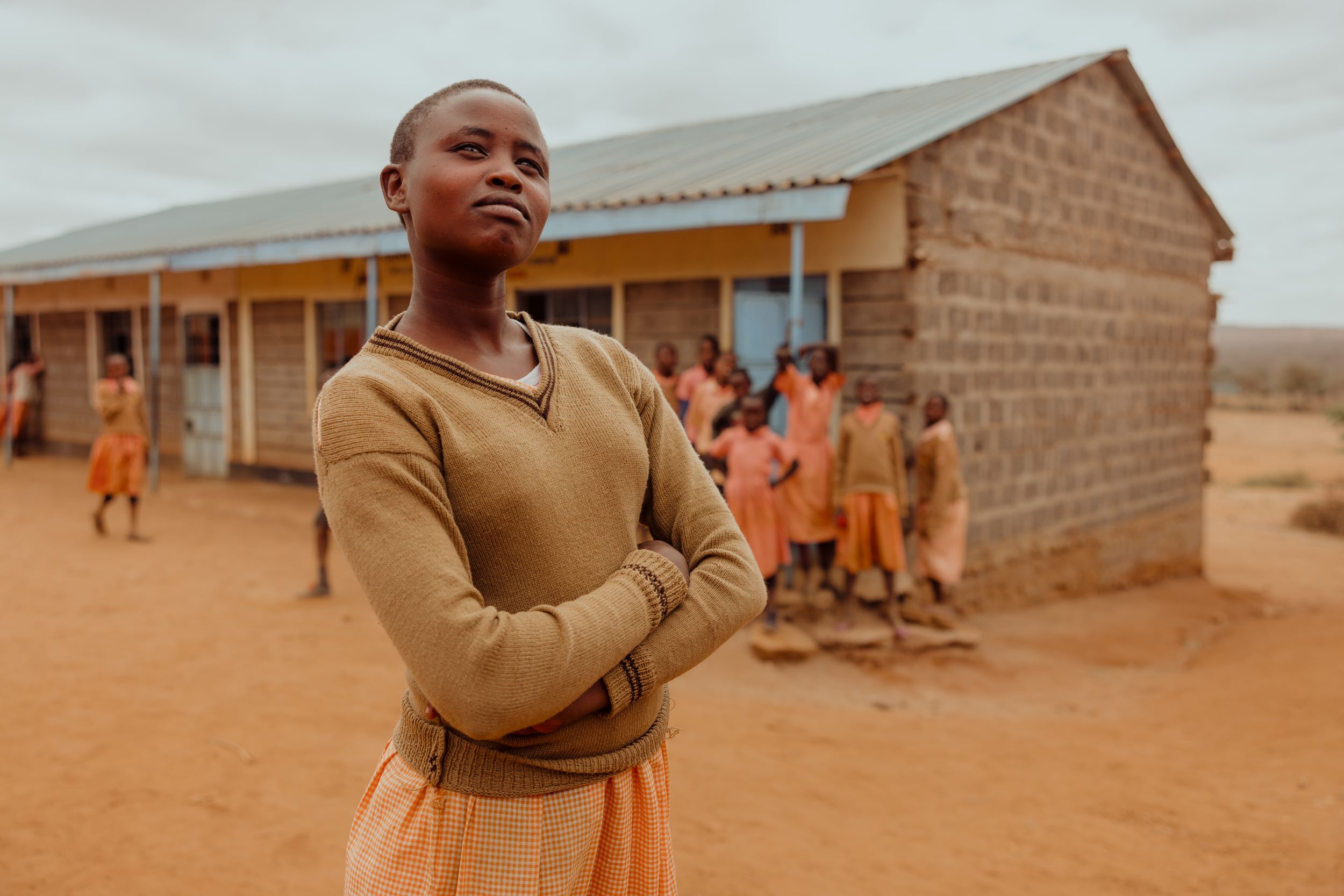 Kenyan girl poses in foreground, behind her school girls playing outside their classroom