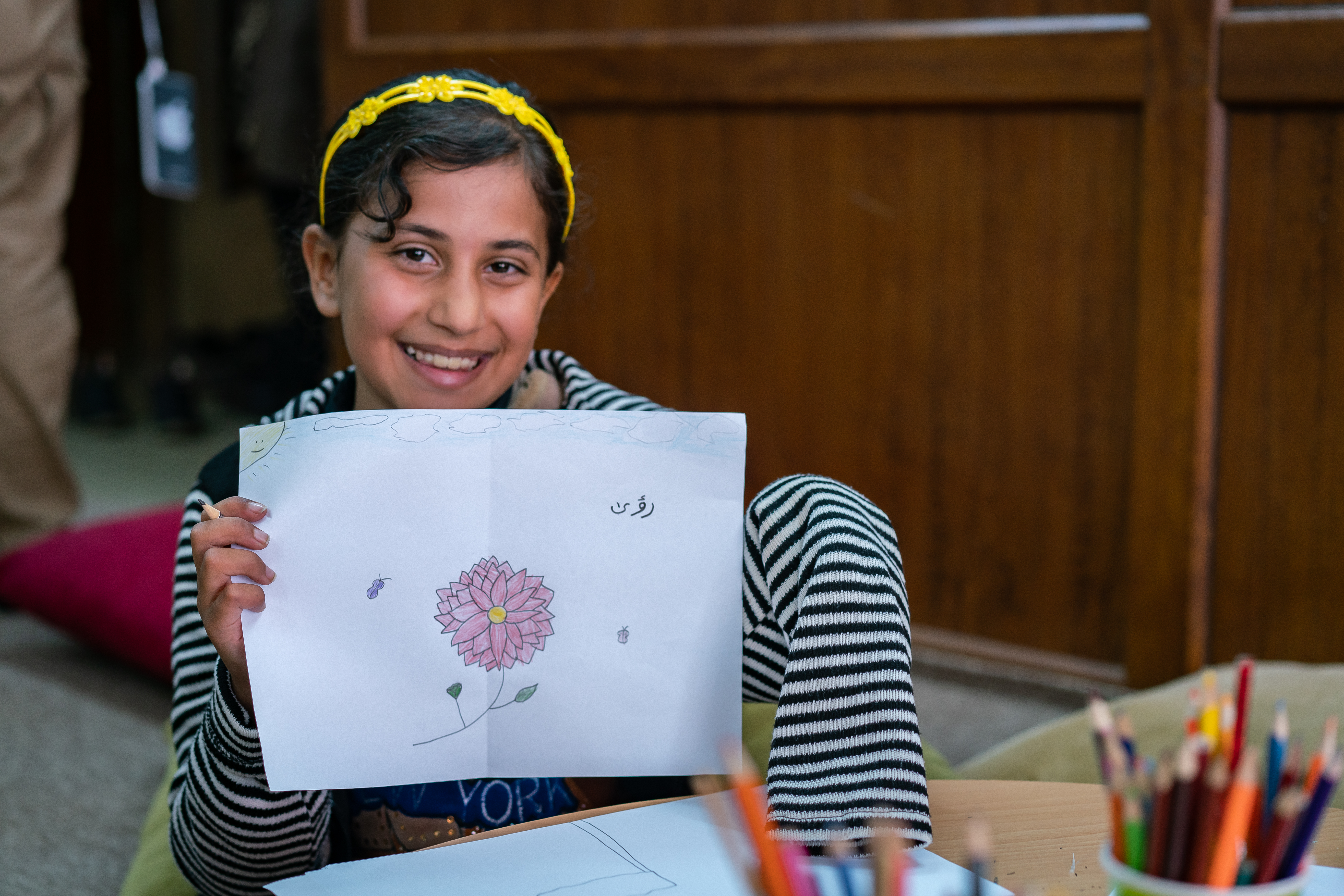 Girl from Iraq smiles as she shows off her artwork, sitting in a room