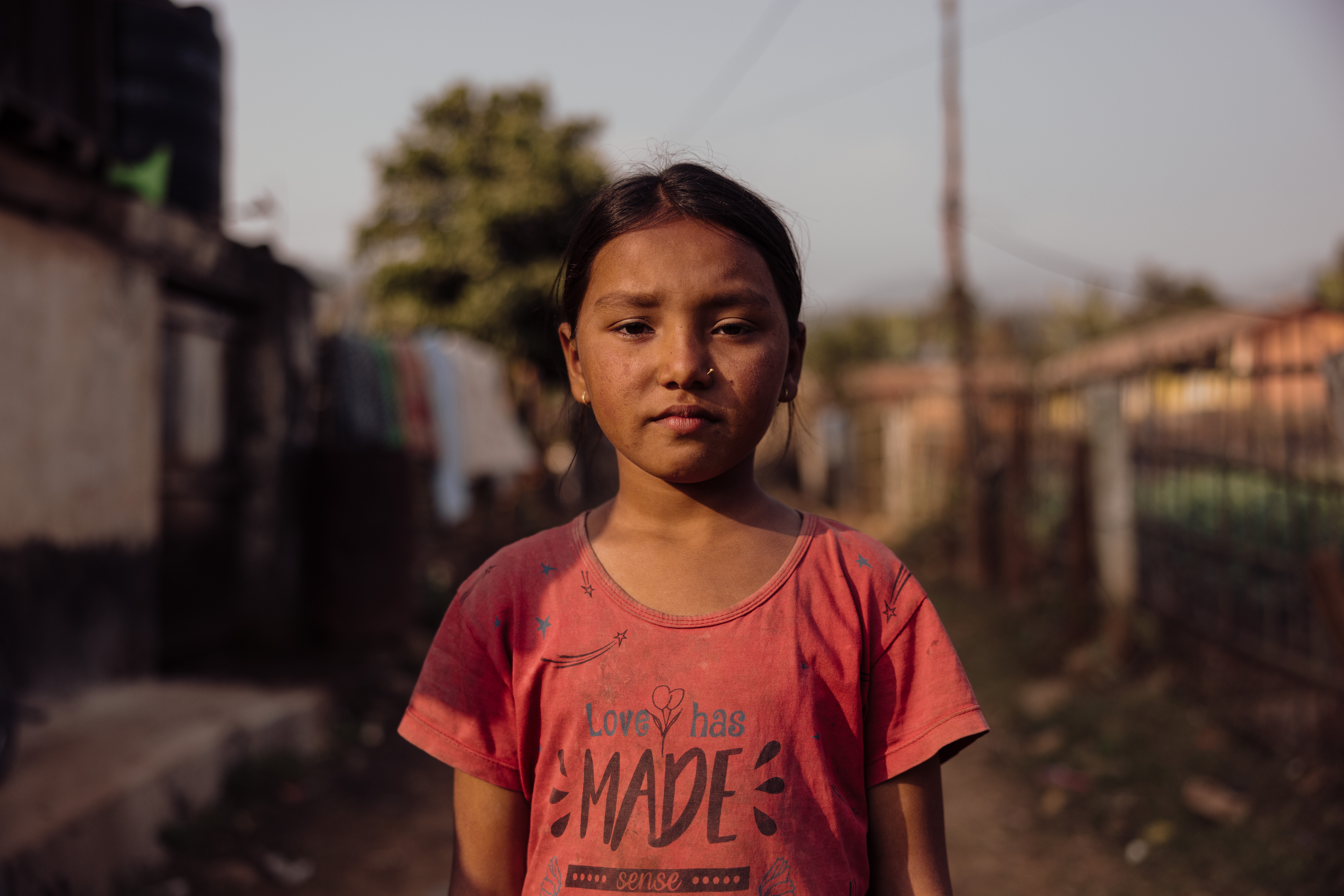 A young girl stands looking into the camera with her home behind hers on hips