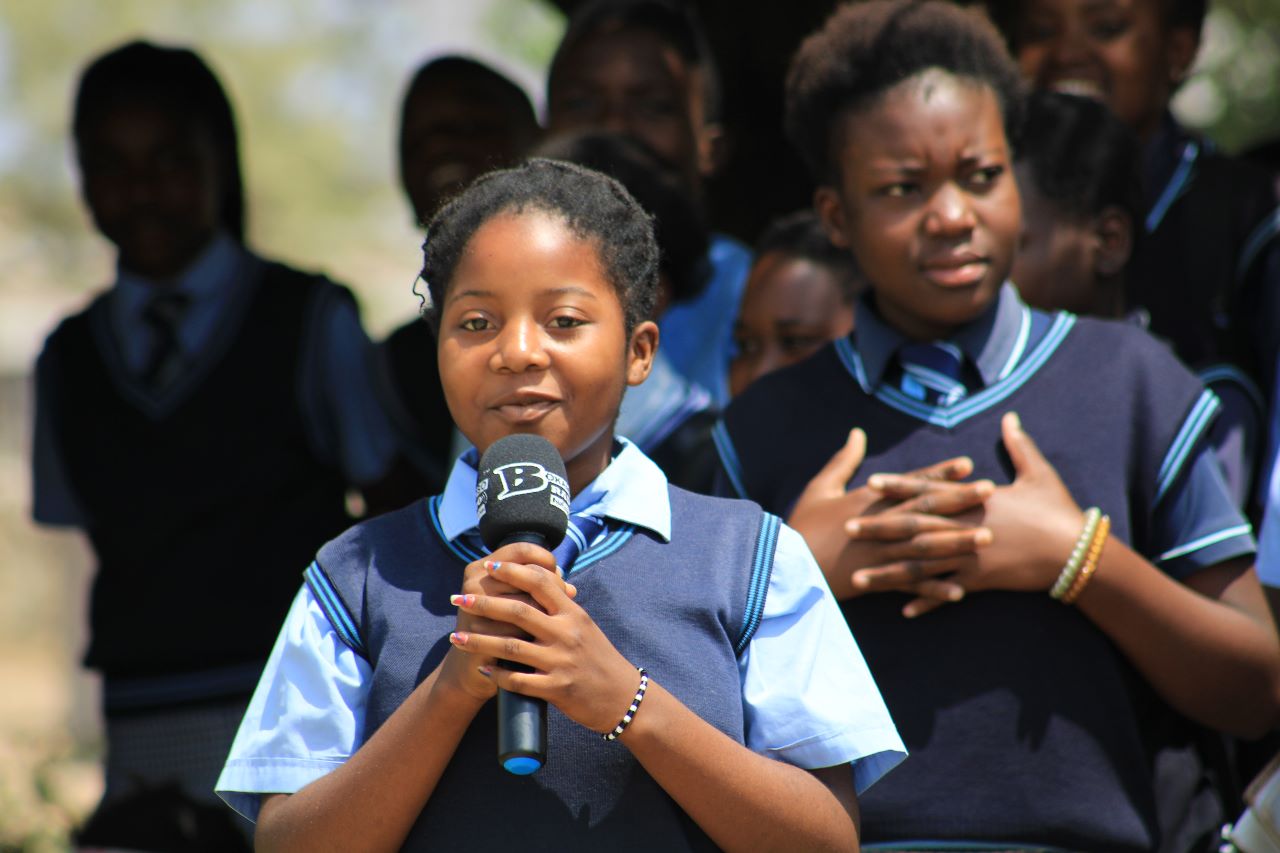 4 children in DRC stand with their hands up and interlaced, standing outside with a few people walking around behind her