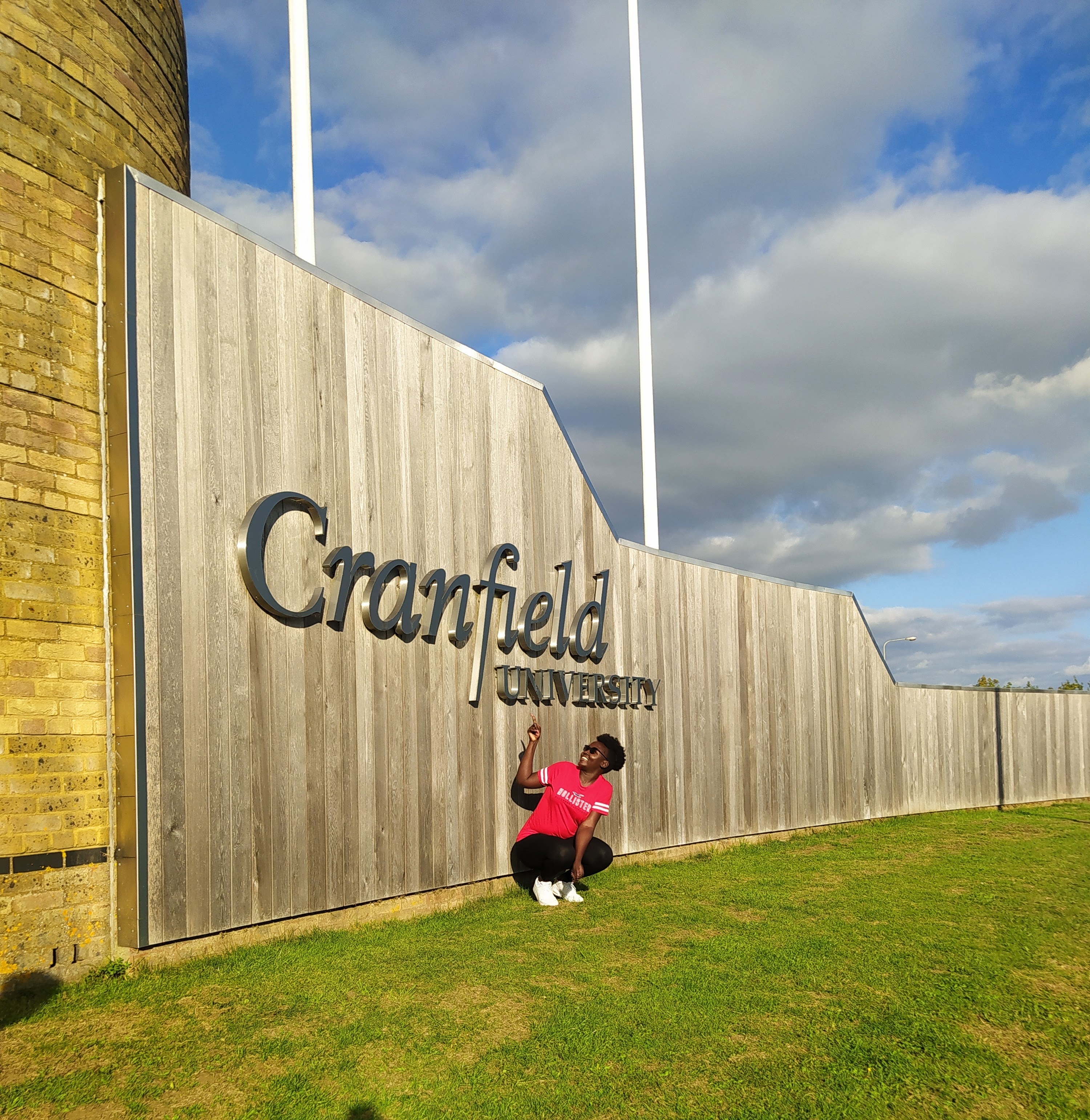 Pauline crouches down and points up at the Cranfield University sign at the campus in Bedfordshire, UK