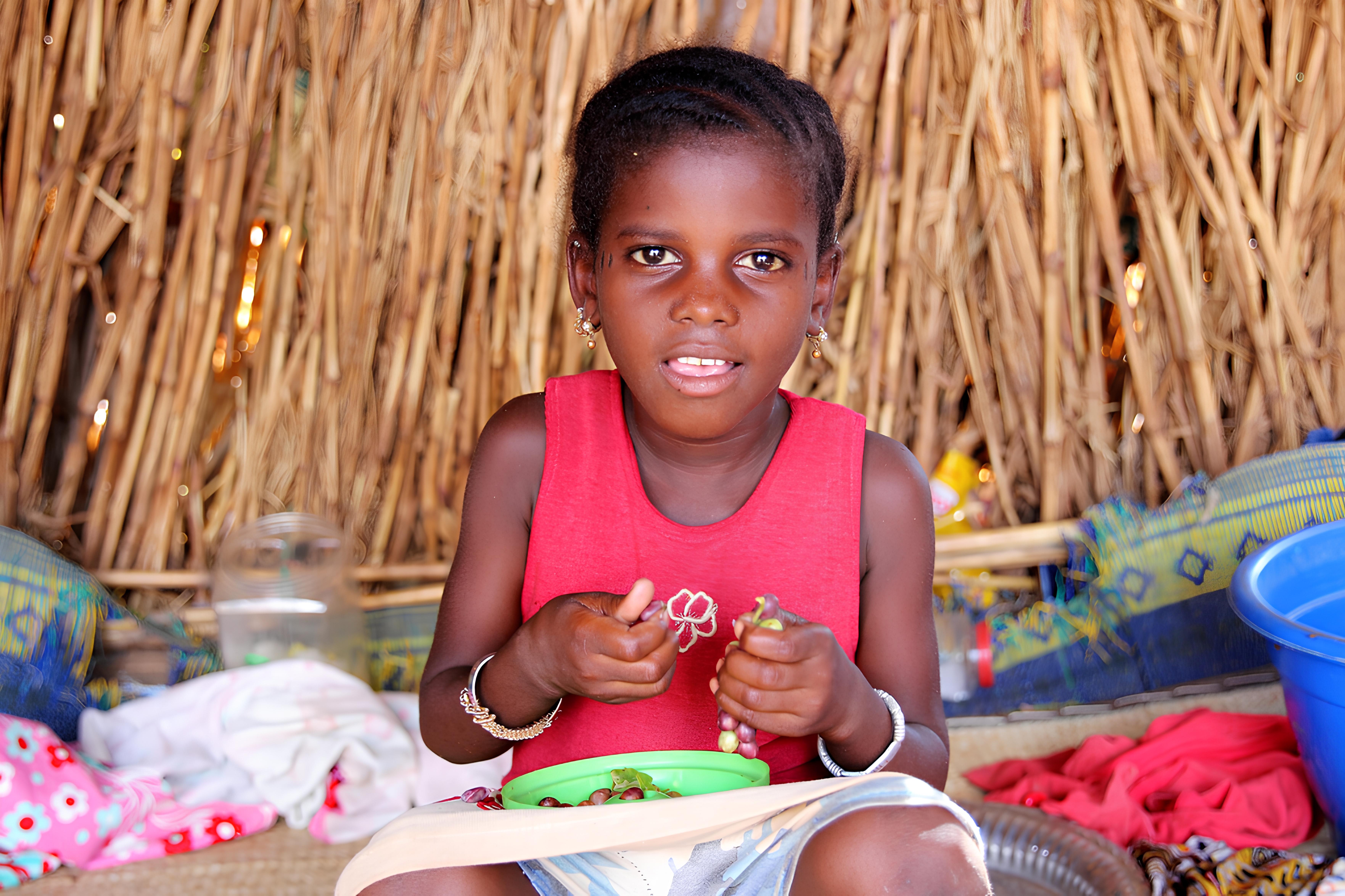 Girl in pink dress sat in front of a thatched background eating grapes and looking directly into the camera