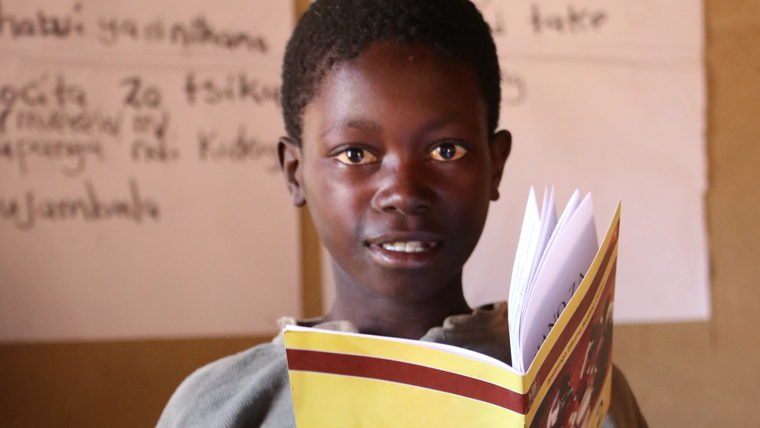 Boy in Zambia looks at the camera as he reads a book in front of a whiteboard on World Book Day