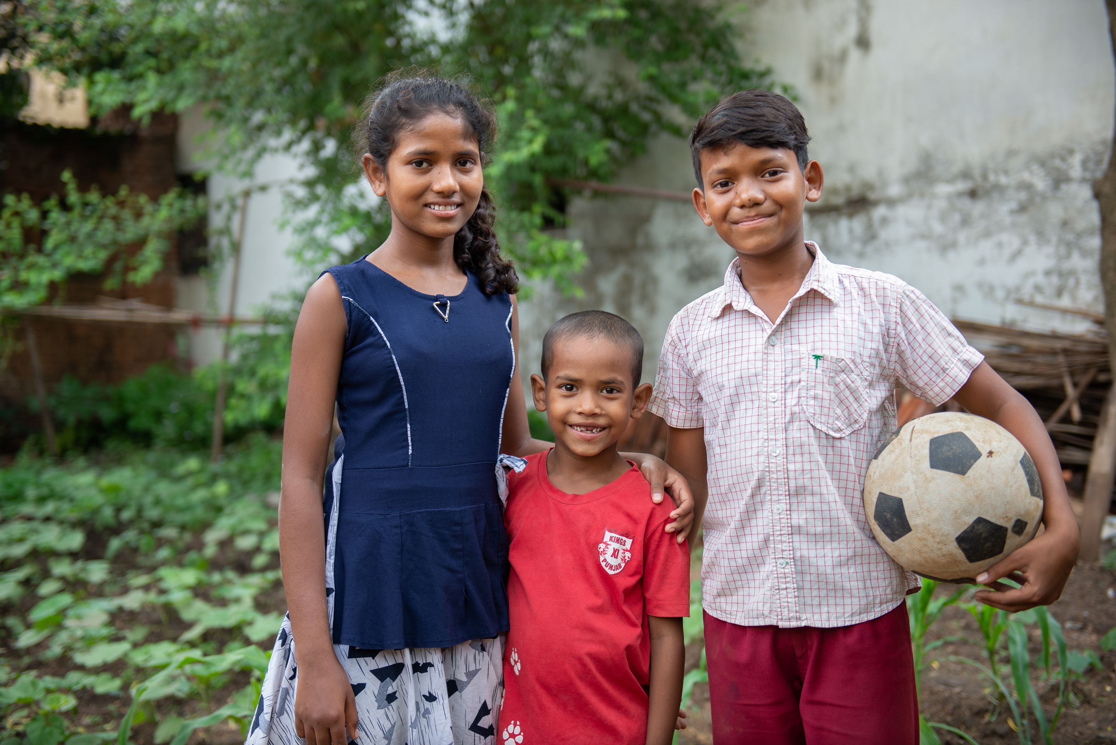 Three children in India smile as they stand together outside, one with a football in his hands, arms around each other