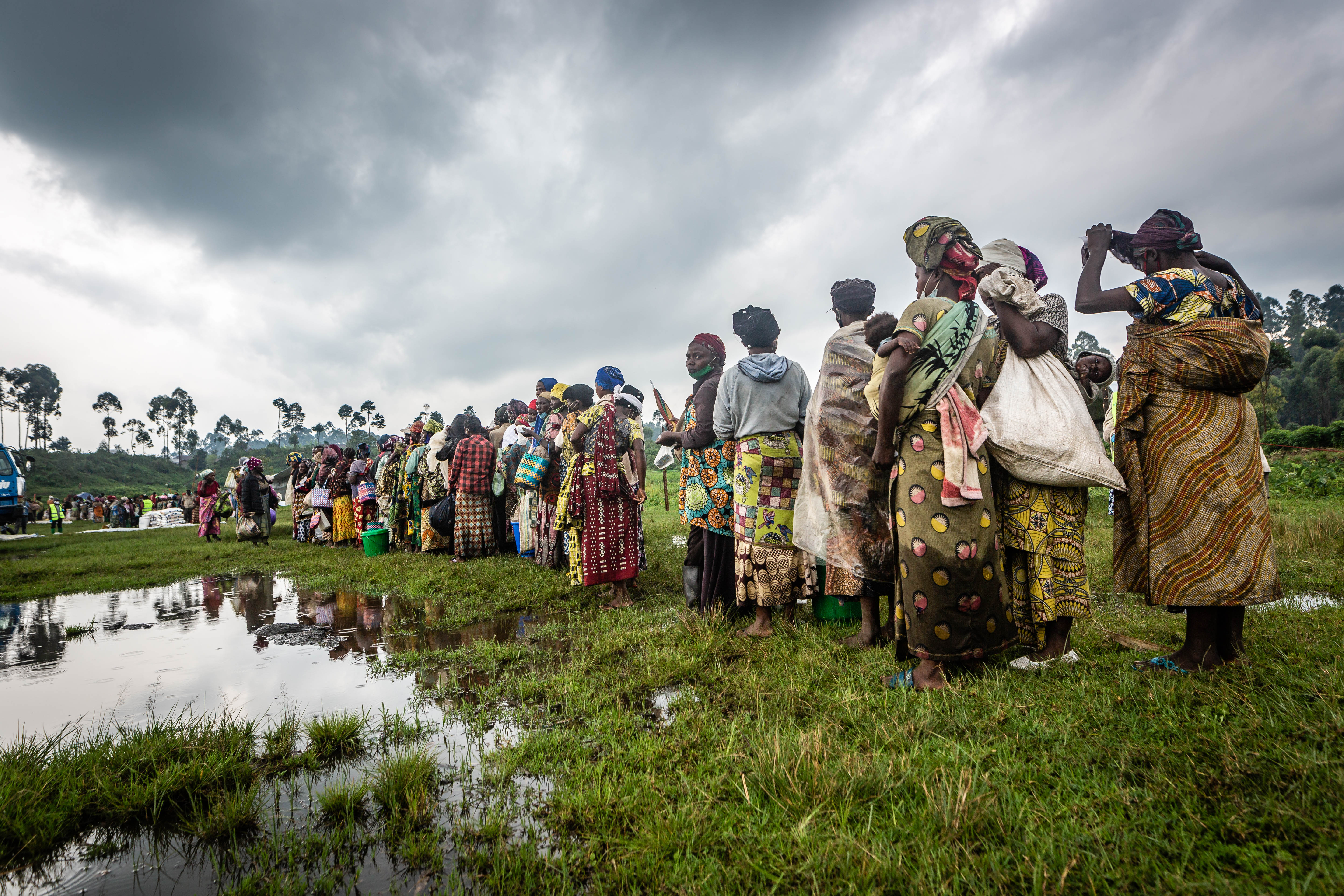 Ques form as people wait for their food packages in the DRC on a cloudy day. The grass is green but muddy from a period of continuous rainfall. 