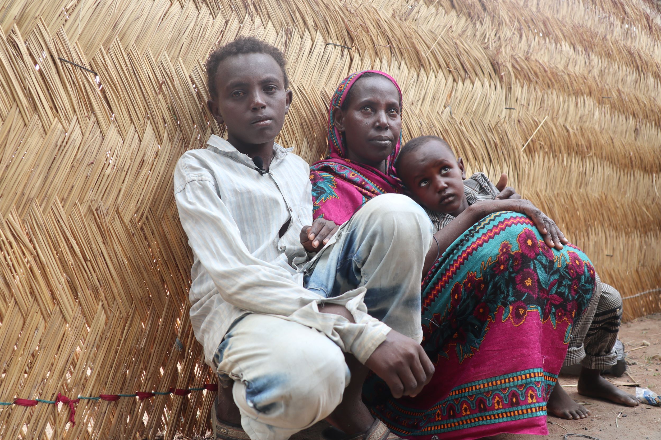 A refugee boy with his mum and sibling sits outside against the woven wall of their shelter in South Sudan
