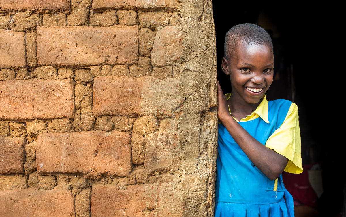 A sponsored child in Kenya wears a blue dress and poses next to a brick wall, smiling.