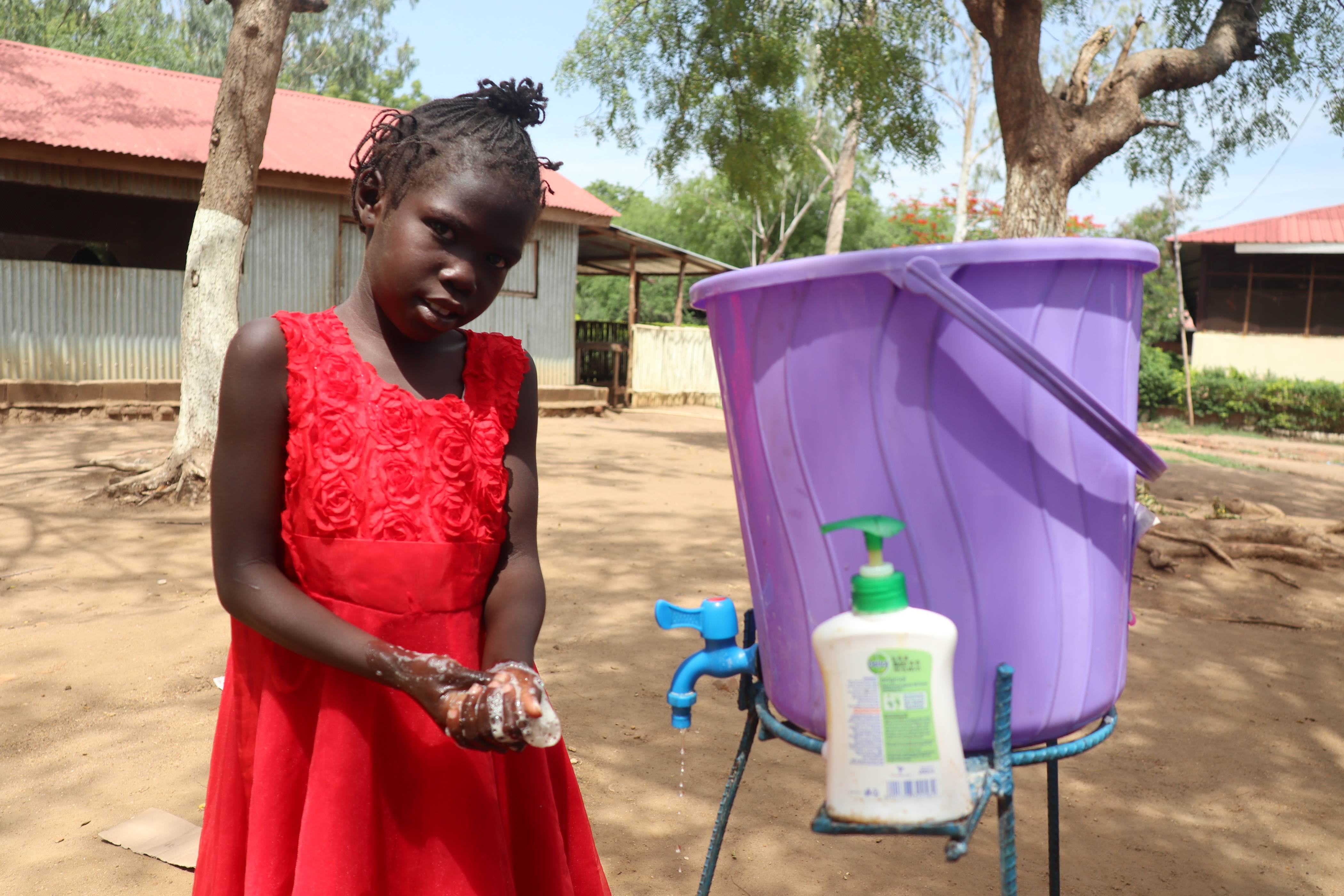 Girl in a red dress in South Sudan washes her hands with water from a purple tub