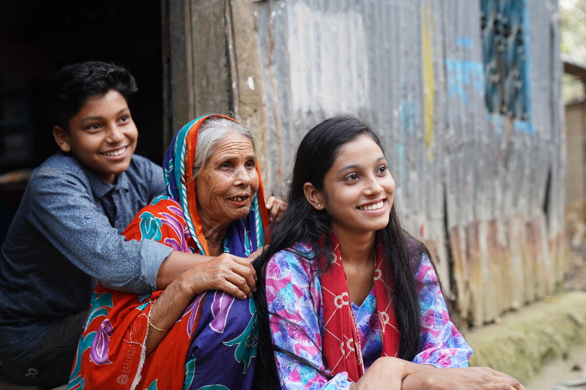 Bangladeshi family sitting in a line outside their home. From left to right, a young boy, grandmother and teenage girl