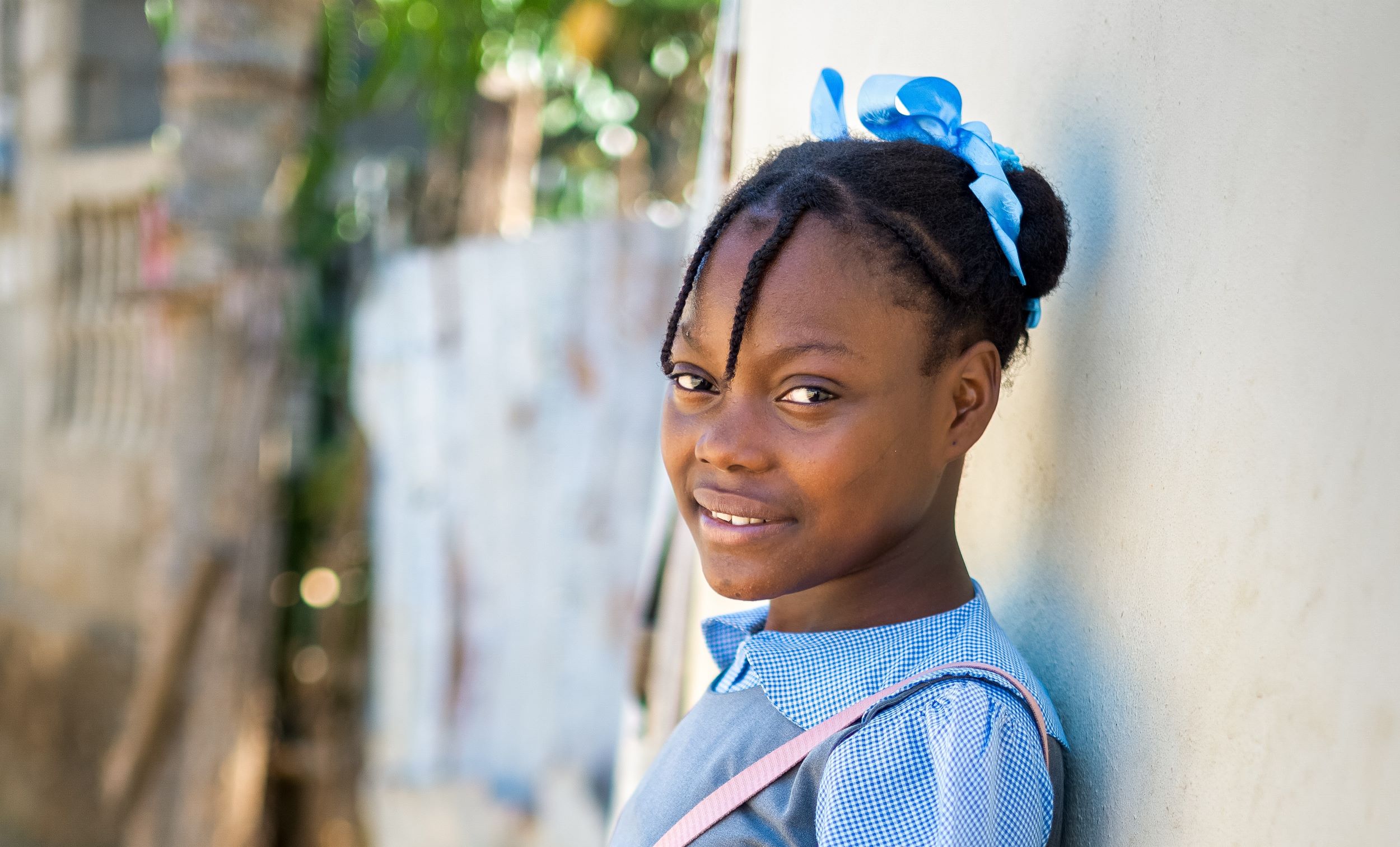 A smiling Haitian school girl holds her pink school bag and leans against a wall