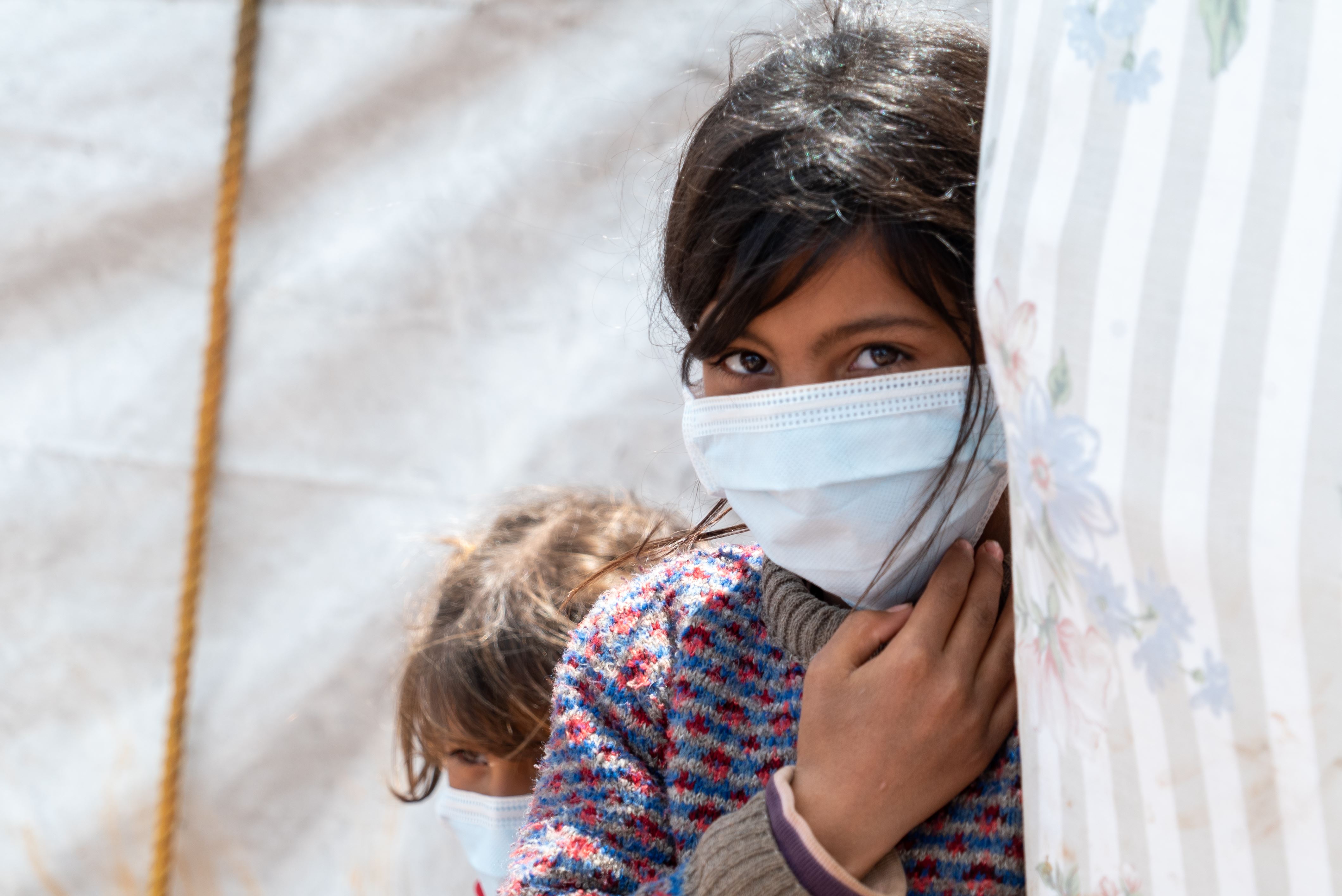 A Syrian girl peeks around the corner of a tent, wearing her face mask
