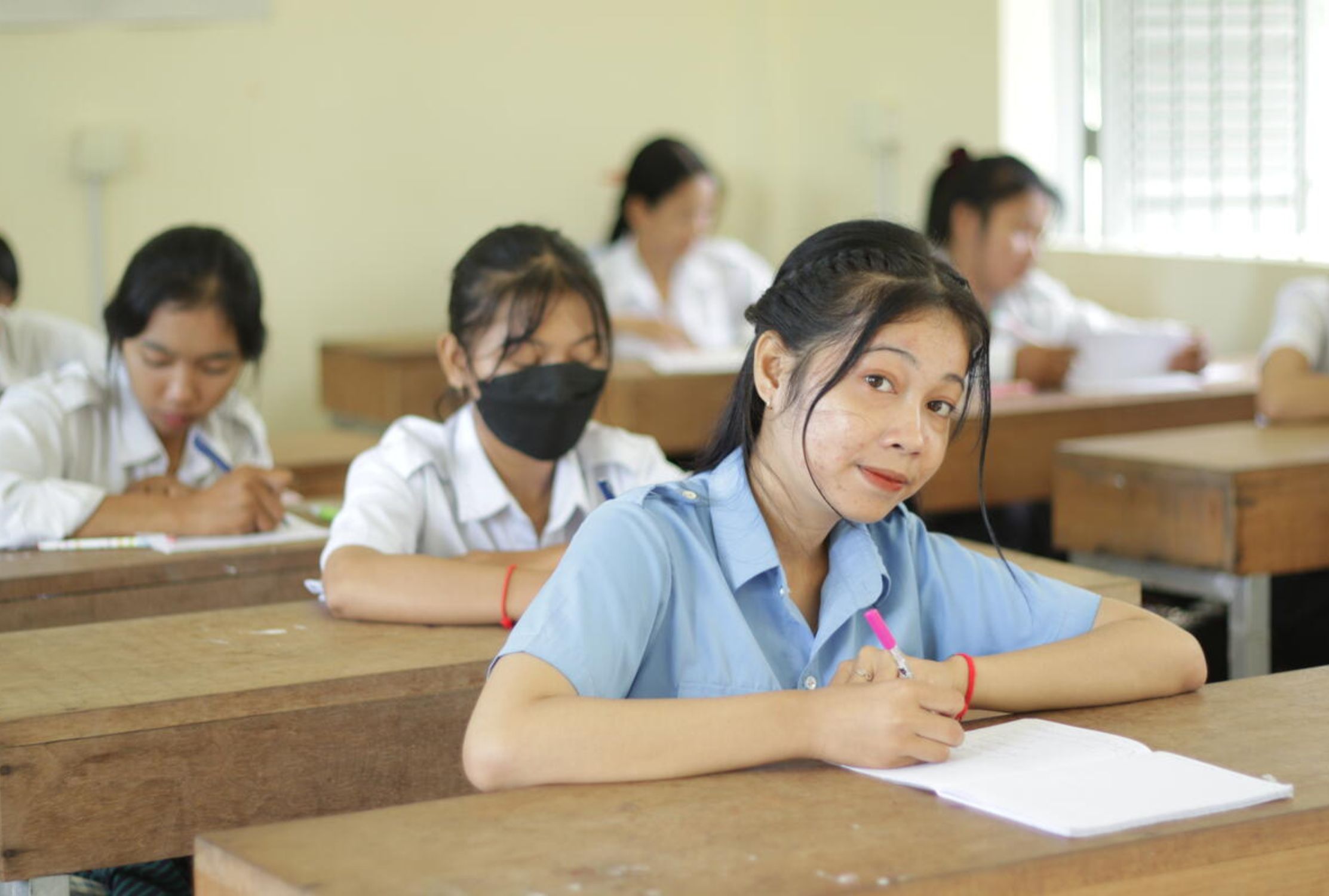 Cambodian school girl sat at a desk in her classroom, smiling at the camera.