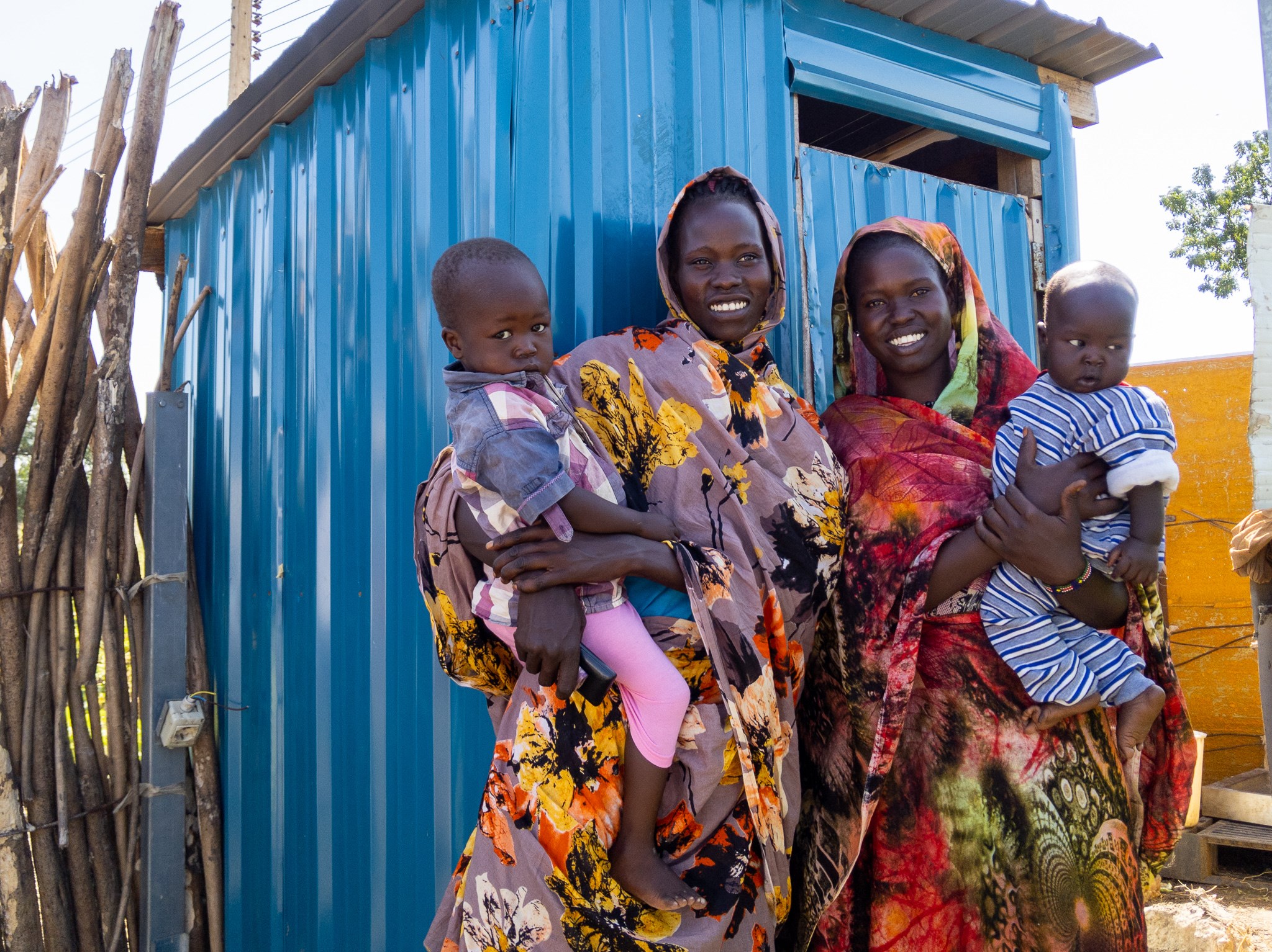 Two mothers from South Sudan holding their babies and standing in front of a toilet smiling to camera