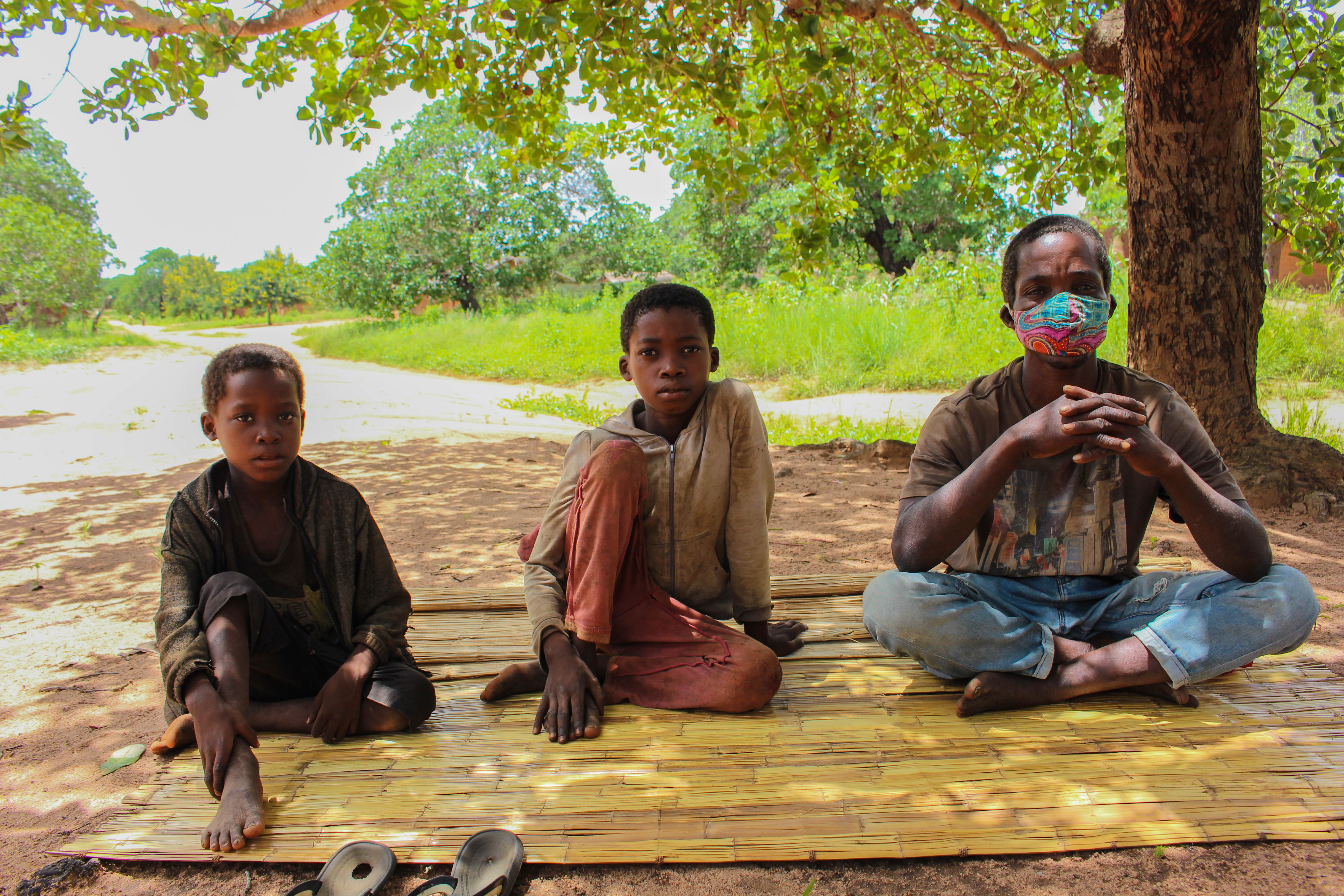 Dércio seated under a tree with his younger brother and member of the community 