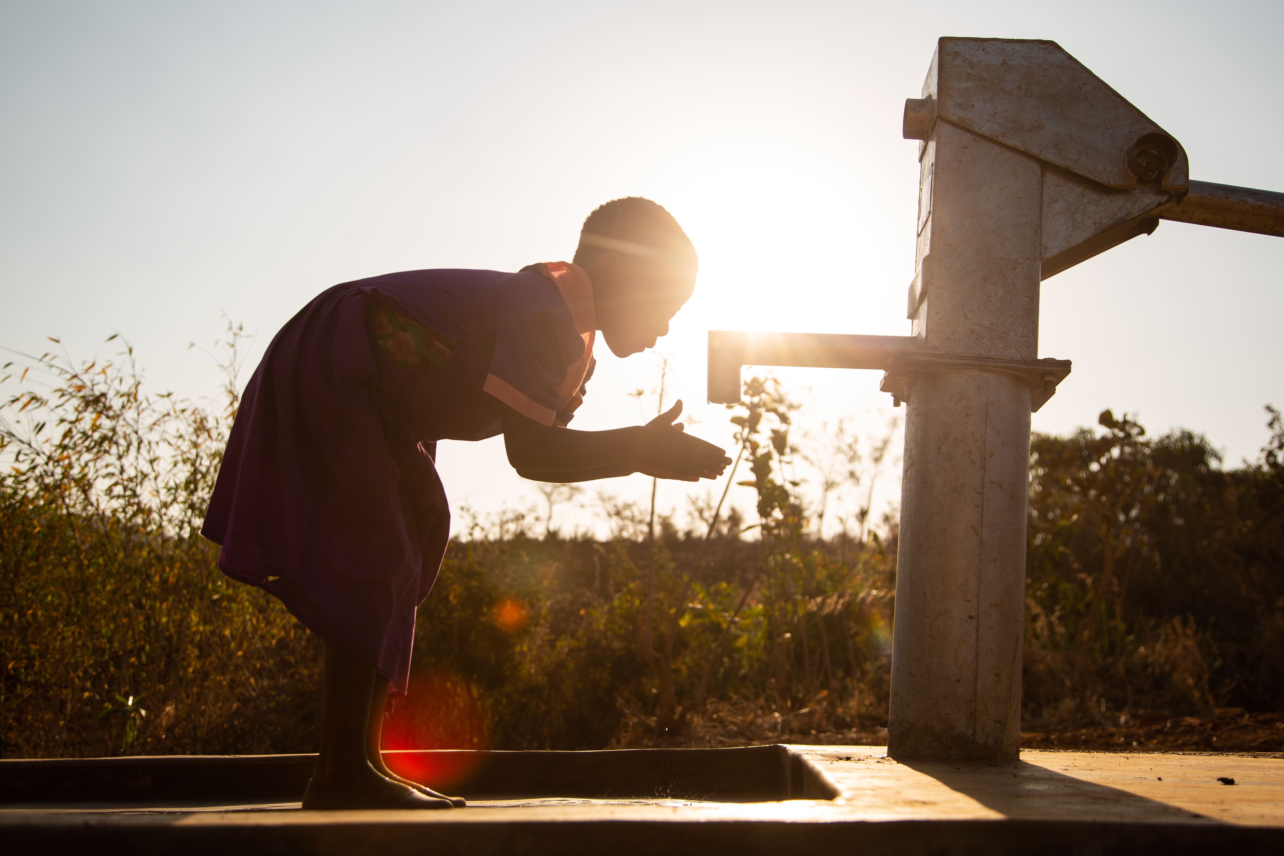 Girl in Malawi bends to clean hands in a clean borehold, silhouetted against the sun