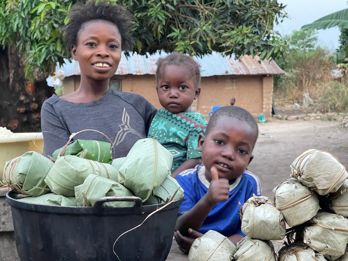 DRC mother standing with her six-year-old son and holding her three-year-old son, behind a large basket of vegetables