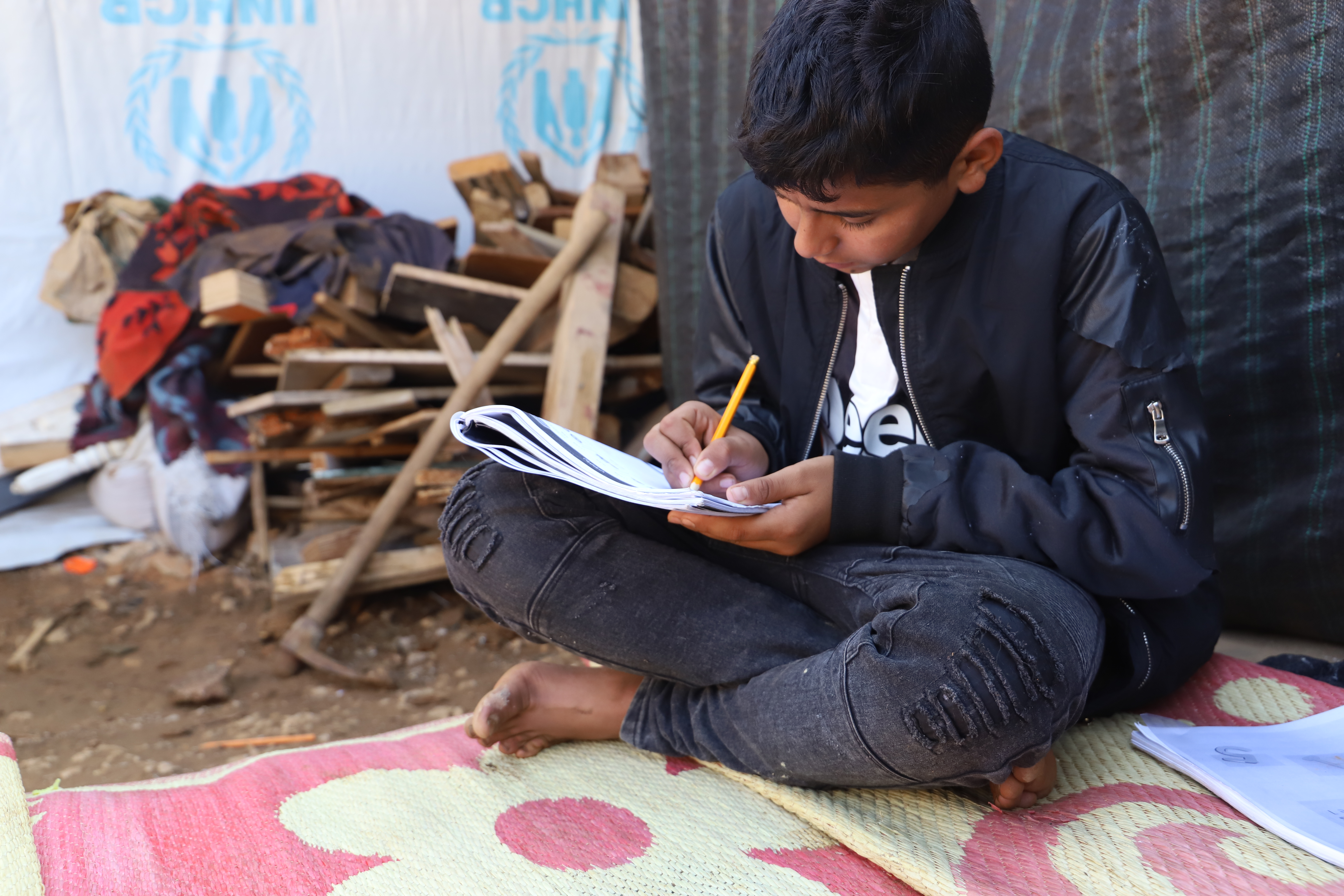 Boy from Syria sitting on the floor at a refugee camp shelter, writing in a notebook