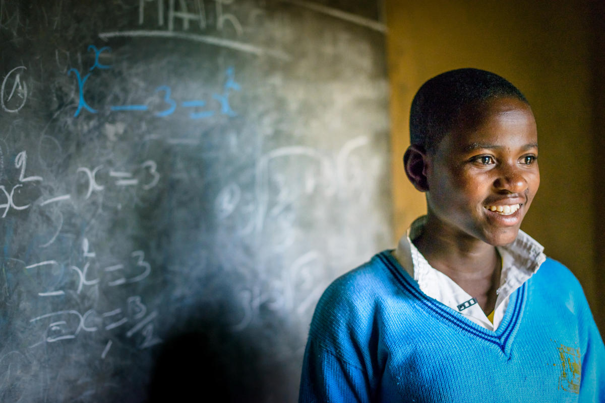 A boy from Rwanda wearing a blue top smiles off to the side of the camera standing with a blackboard behind him