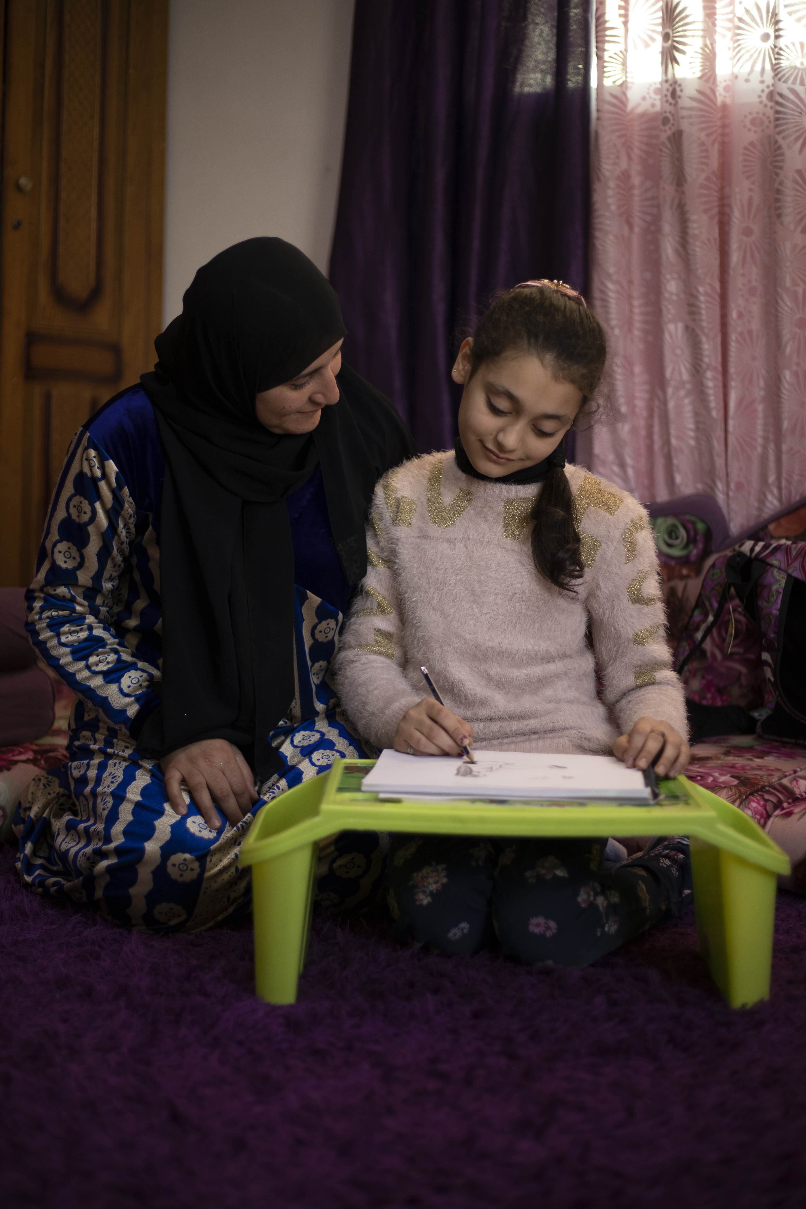 Ten year old Rahma sitting down at a table with her mum showing her the drawings she's created