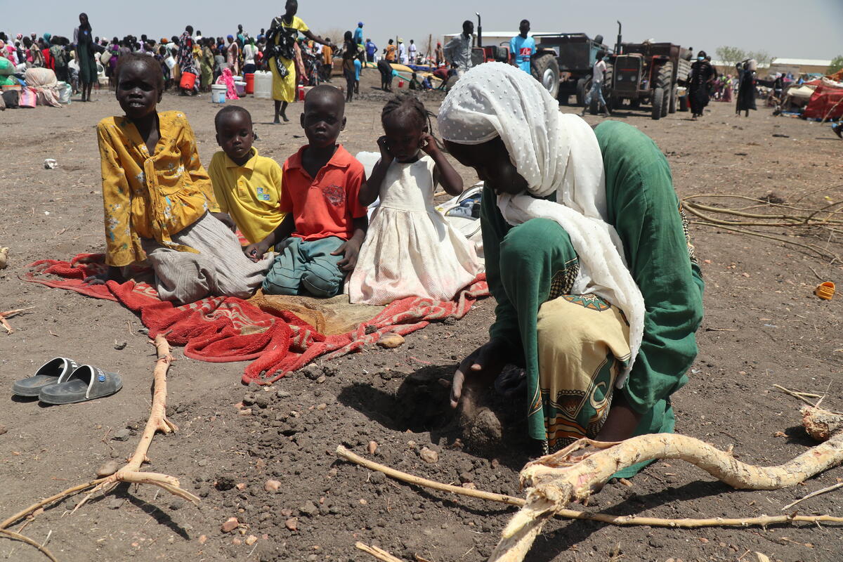 Sudanese refugee family sat on the floor in a displacement camp. Behind them are crowds of other displaced people.