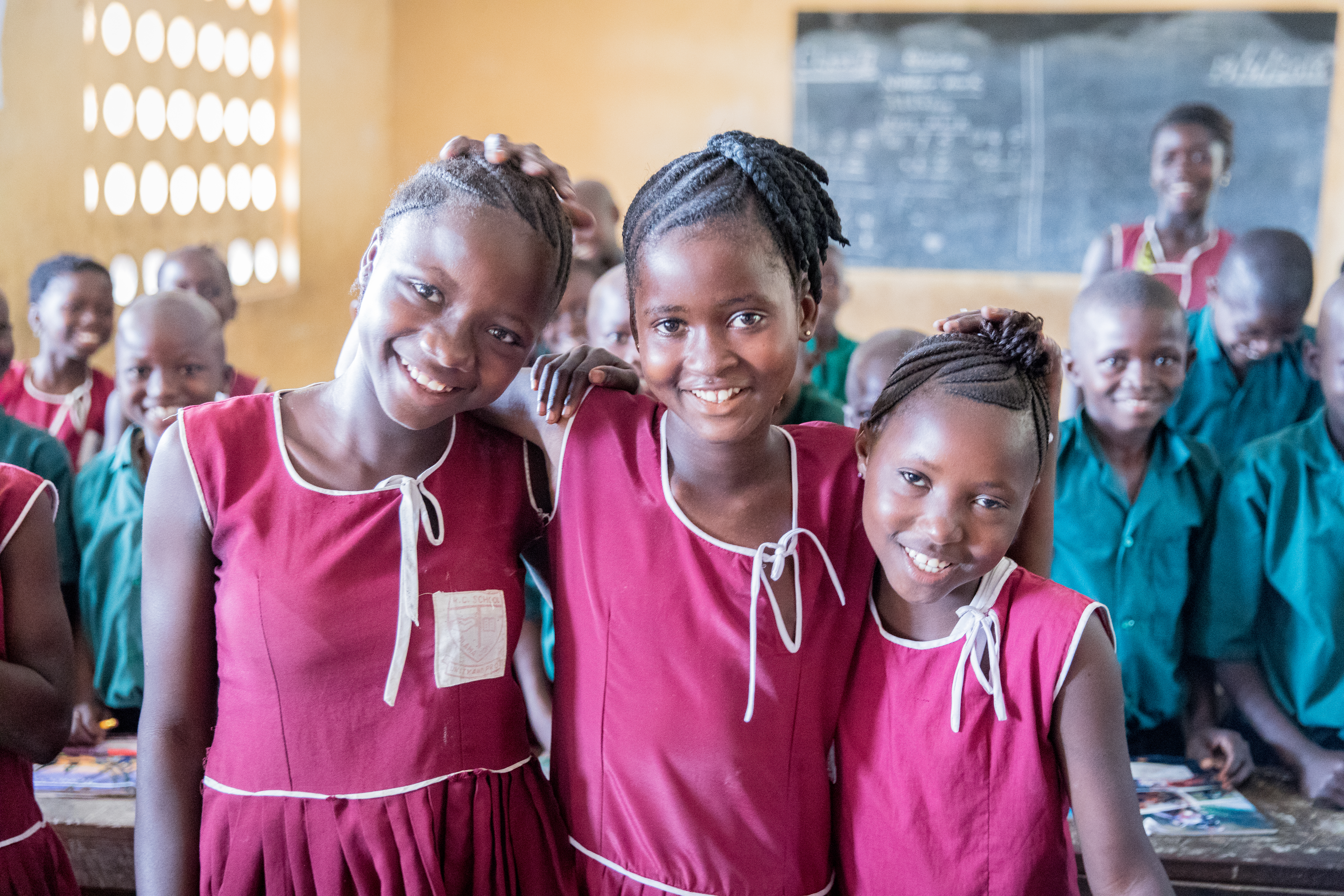 Three girls in red dress school uniforms smile as they stand in their classroom in Sierra Leone
