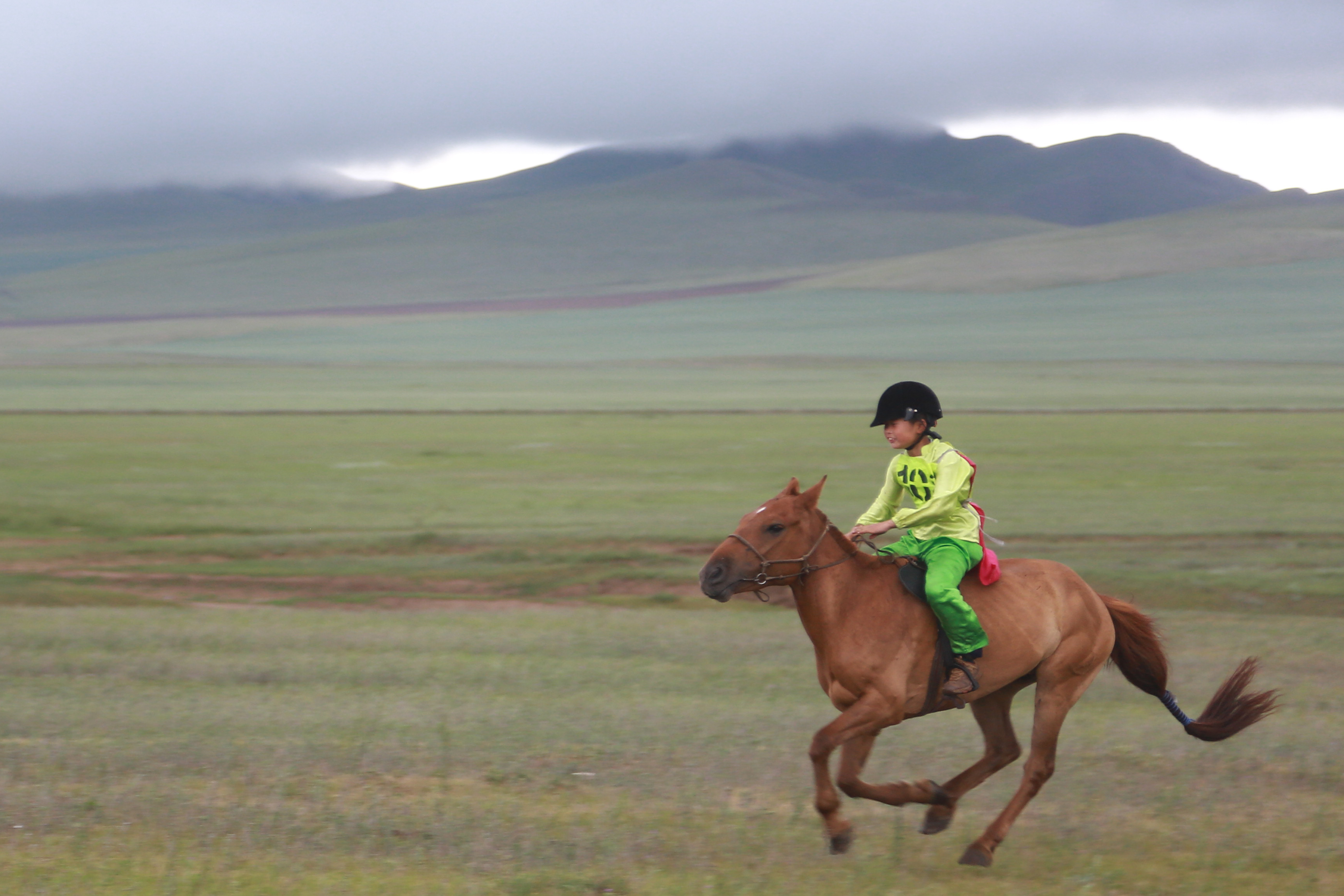 A boy rides his horse across open plains, with mountains and low cloud behind