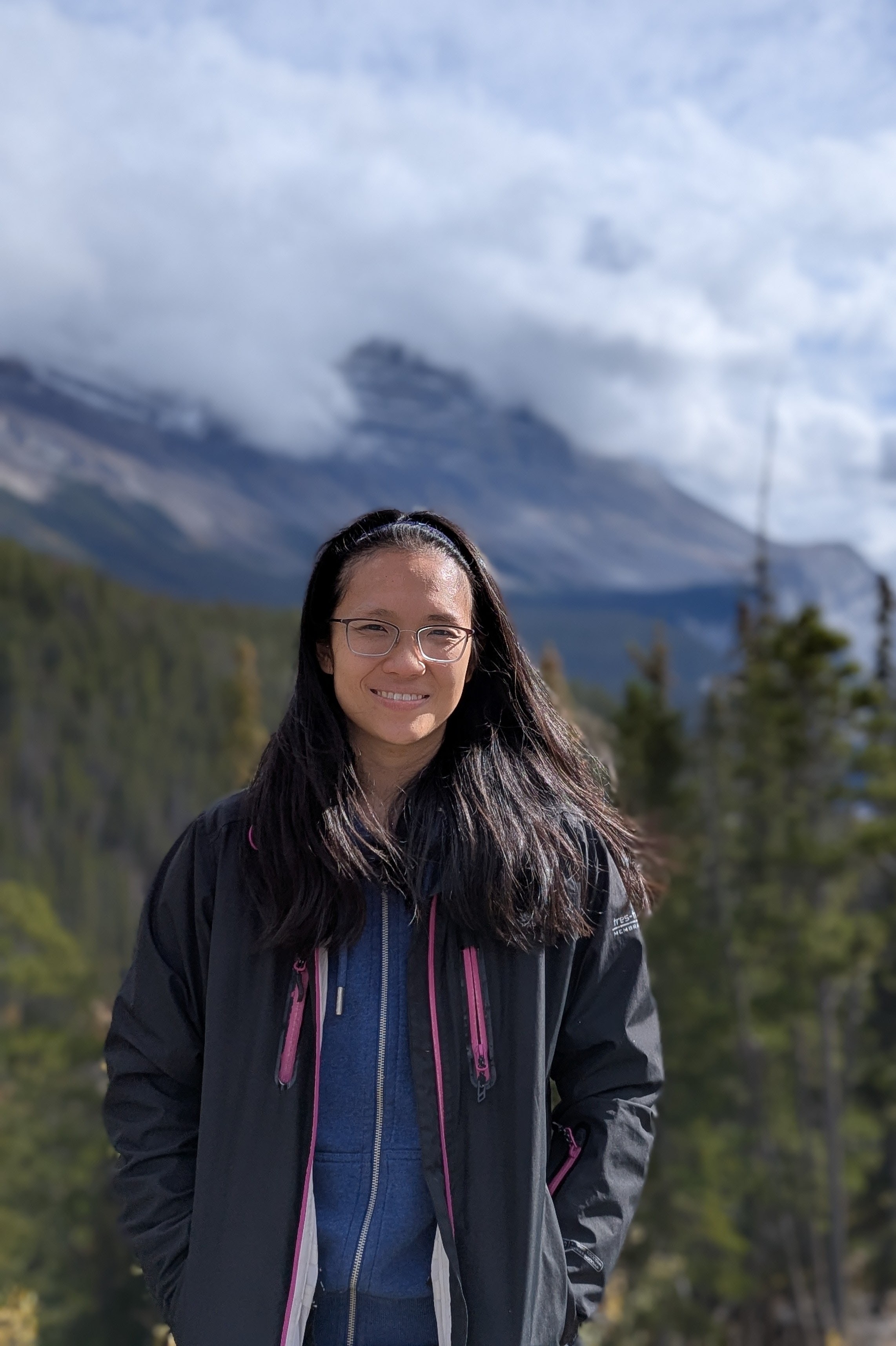 Pei smiles to camera. There's a forest and mountain range in the background.