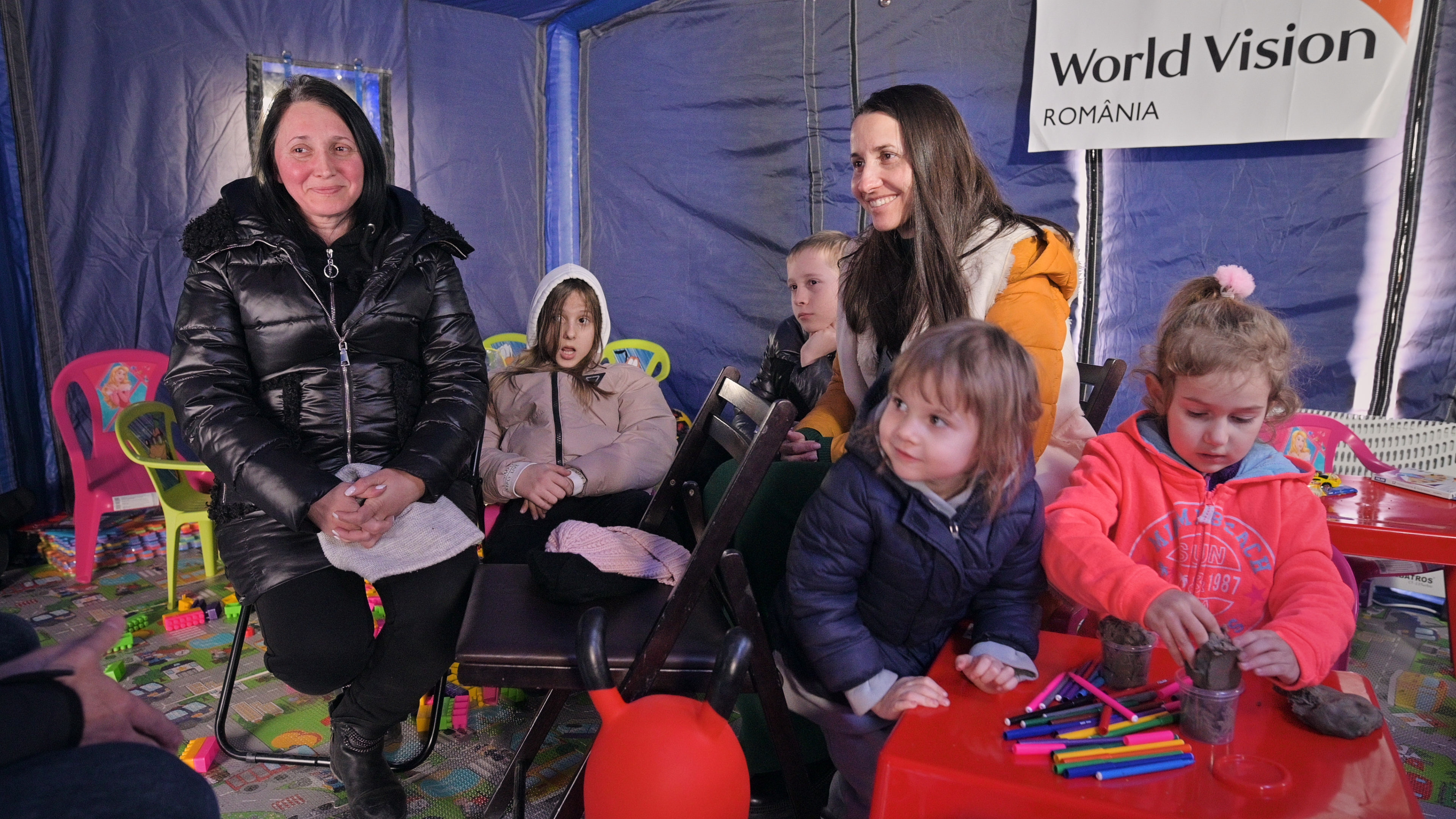 A family in a child friendly space in Romania. Two of the children are playing with clay and playdough while the other four are smiling.