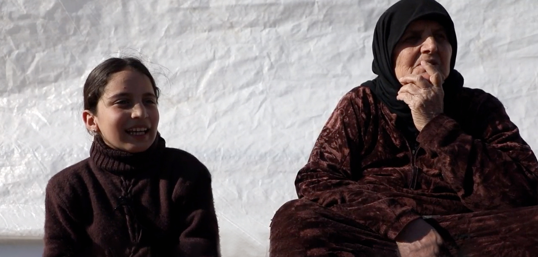 Older lady and granddaughter sit outside their tent in a camp in Syria