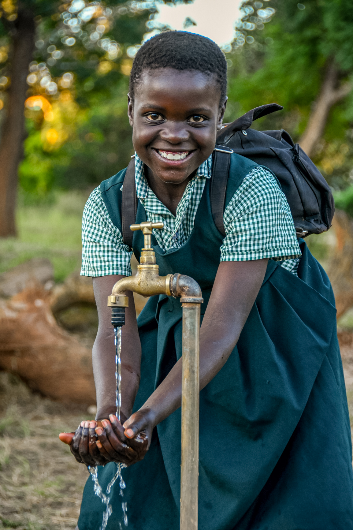 A girl in school uniform smiles and washes her hands in the clean water from a standing tap