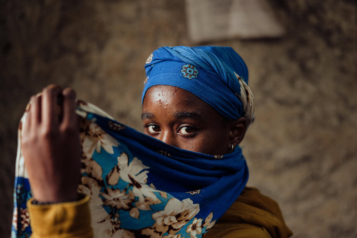 A young girl shields her face with a blue scarf, just her eyes are visible