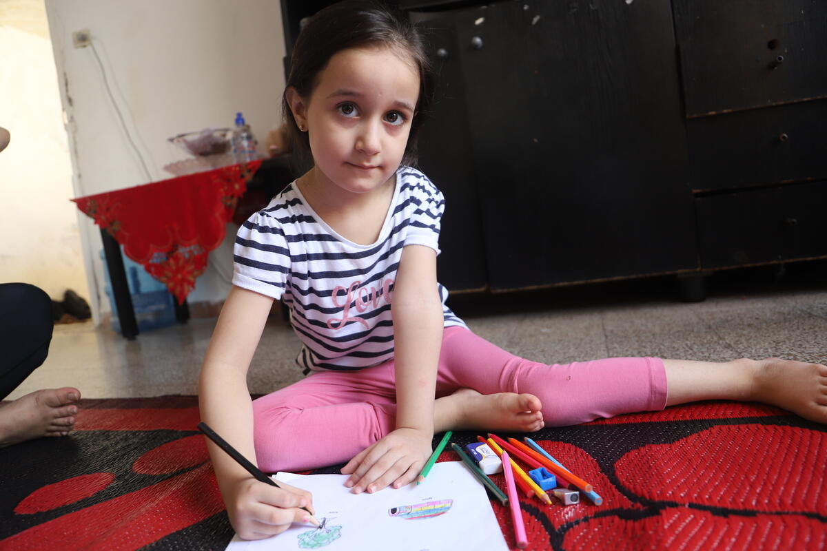 A young girl, 5-year-old Aline,  sits on the floor writing in her exercise book