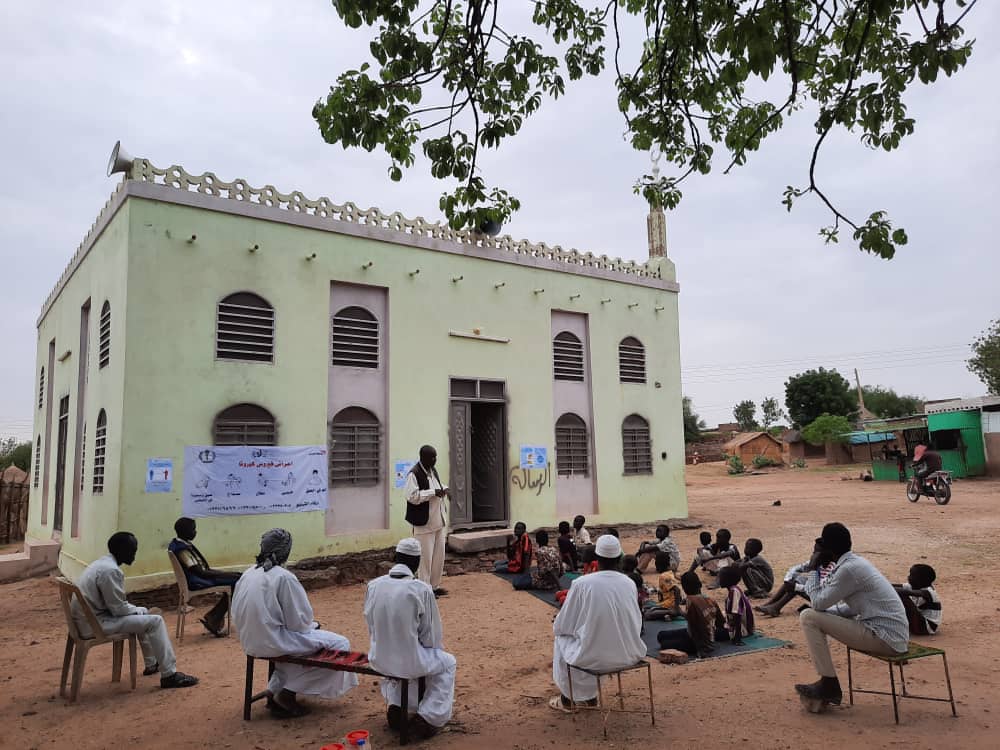 Faith leader stands outside a church in Sudan telling people about good hygiene practices for COVID-19
