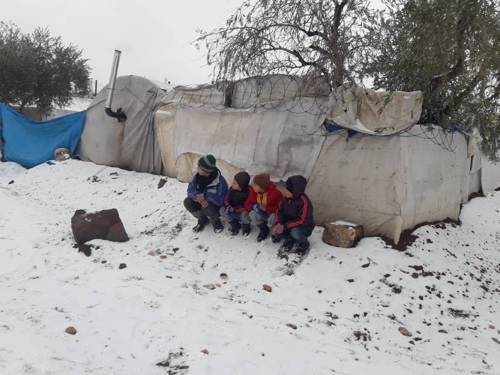 A group of refugee children huddle together next to a tent, in the snow in a camp in Syria