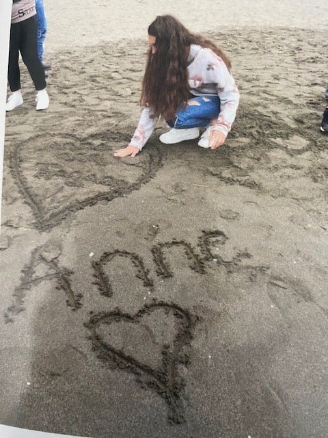 A girl writes Anne's name in the sand with a heart.