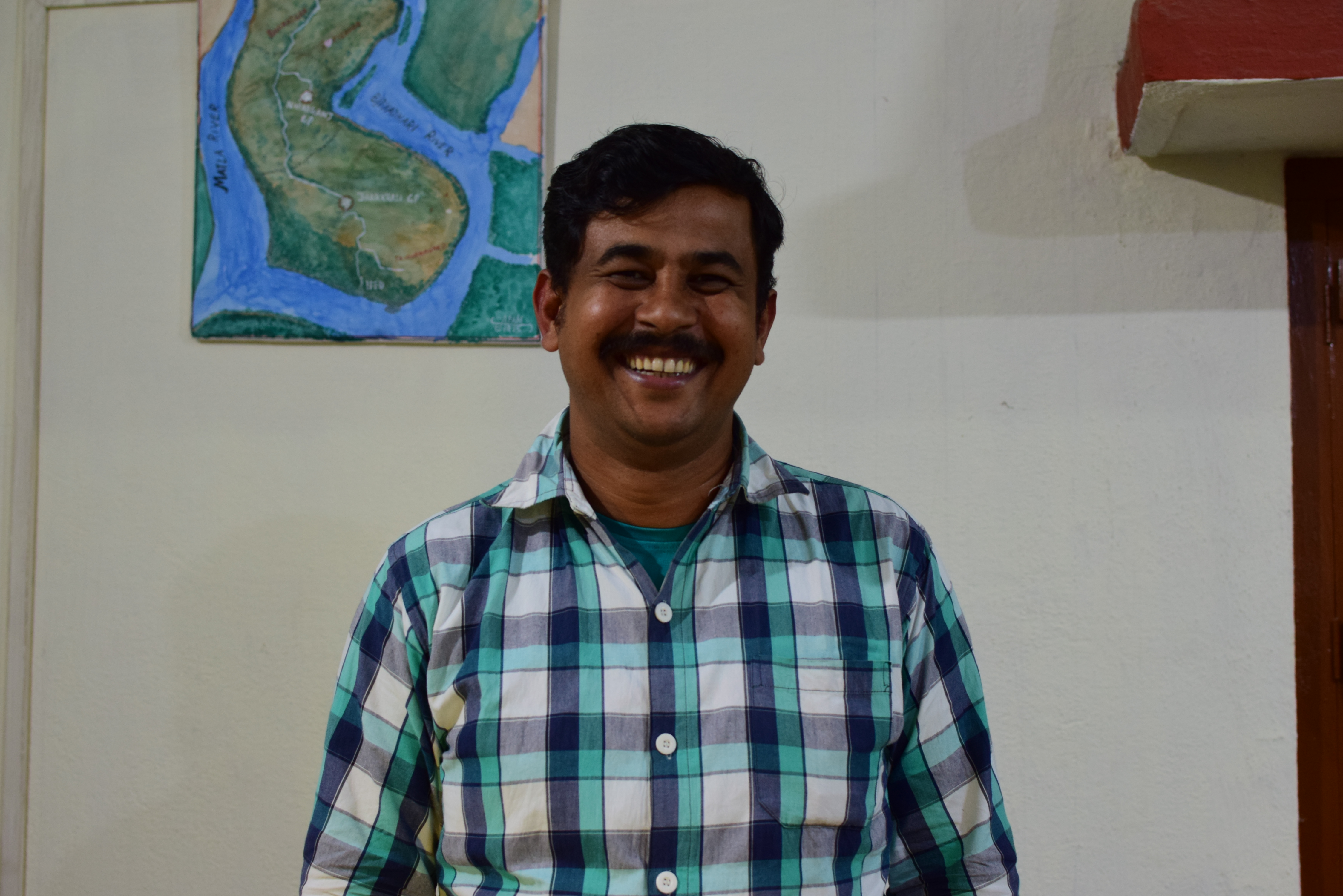 Man in India wears a checkered shirt and smiles as he stands in a room