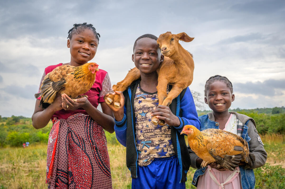 Three siblings pose with livestock gifted to them. Two have hens and one has a goat over his head.