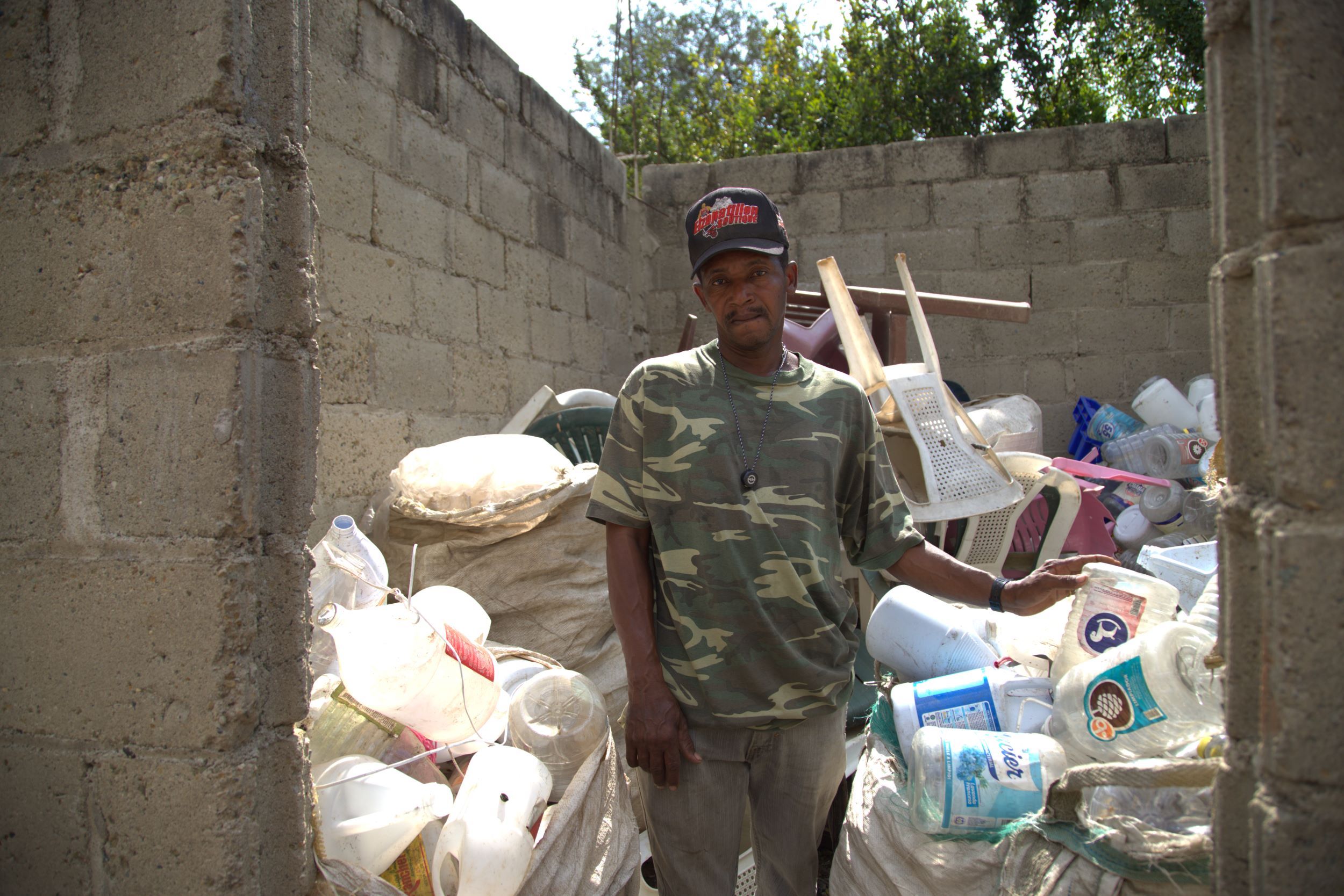 A man from Haiti stands before a mountain of plastic and other recycling he’s collected to sell