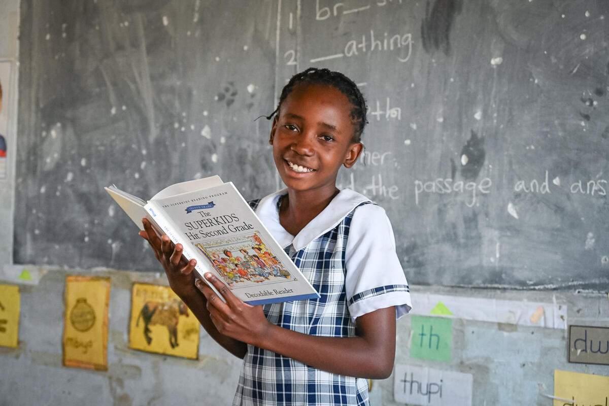 Zambian schoolgirl holding a book and smiling, classroom chalkboard behind her