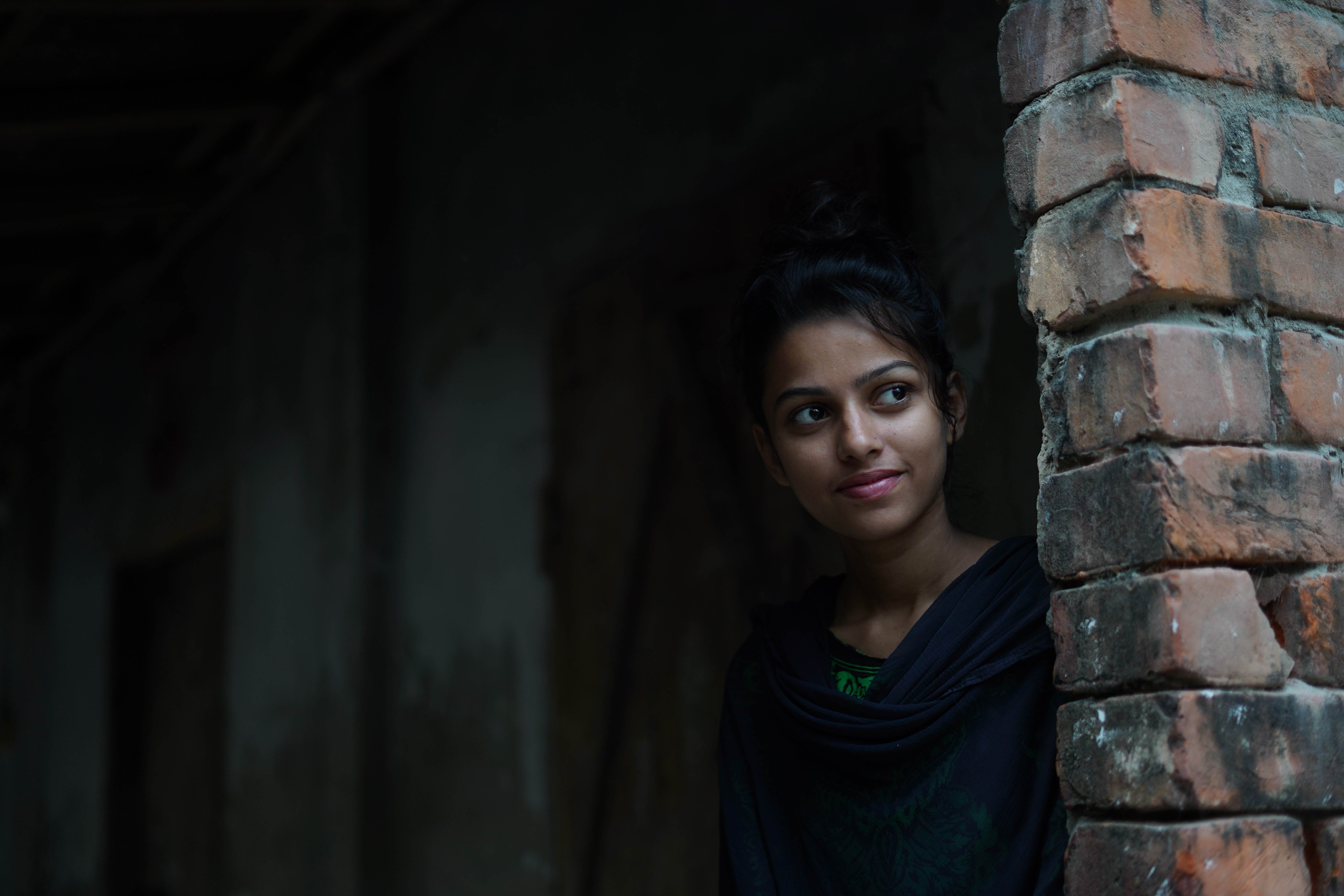 A young woman stands in shadows, peering out from around a brick wall
