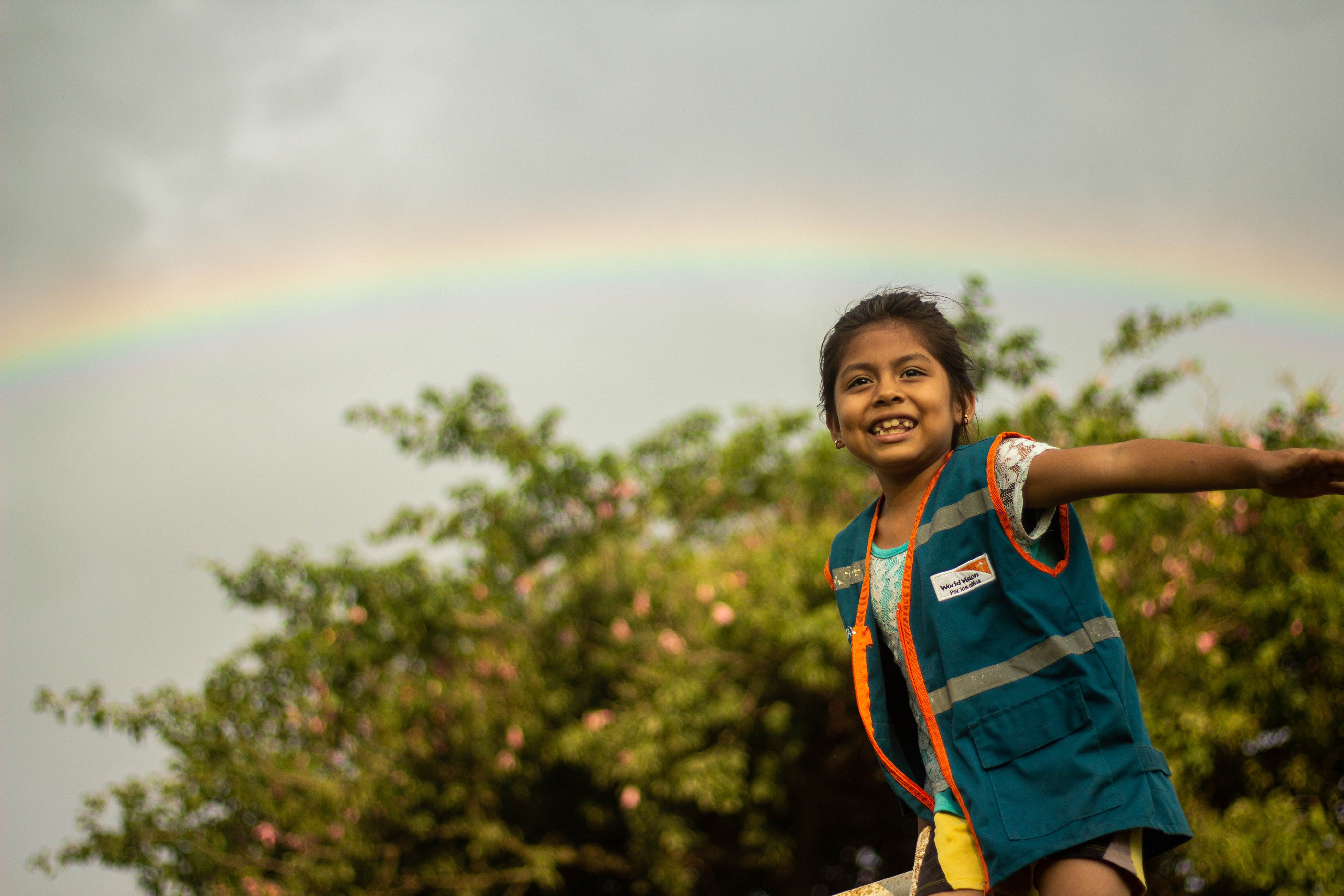 A child smiles at the camera as she pretends to fly in front of a rainbow in Bolivia 