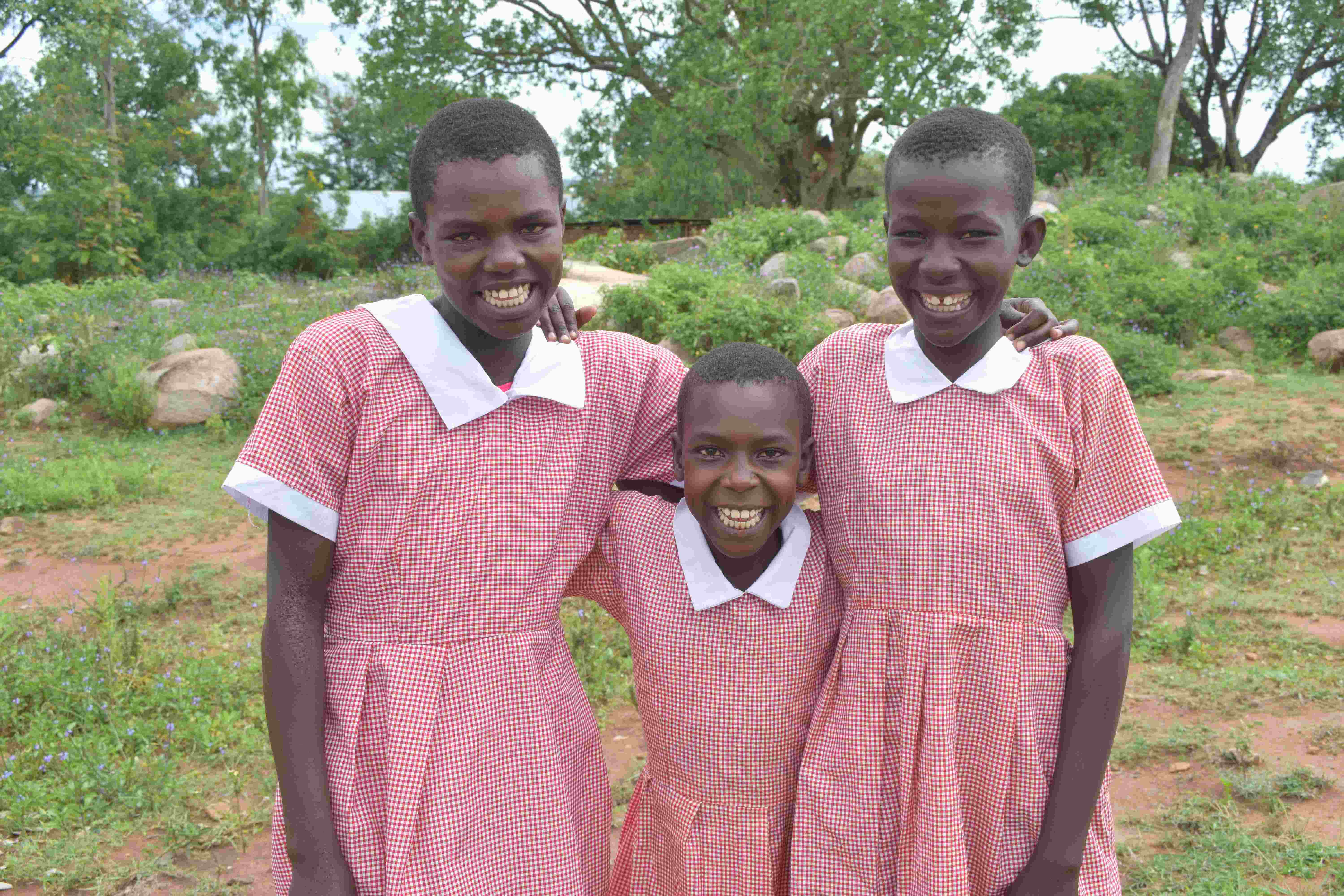 Three Kenyan girls wear pink dresses for school, and stand with their arms around each other, out in a field
