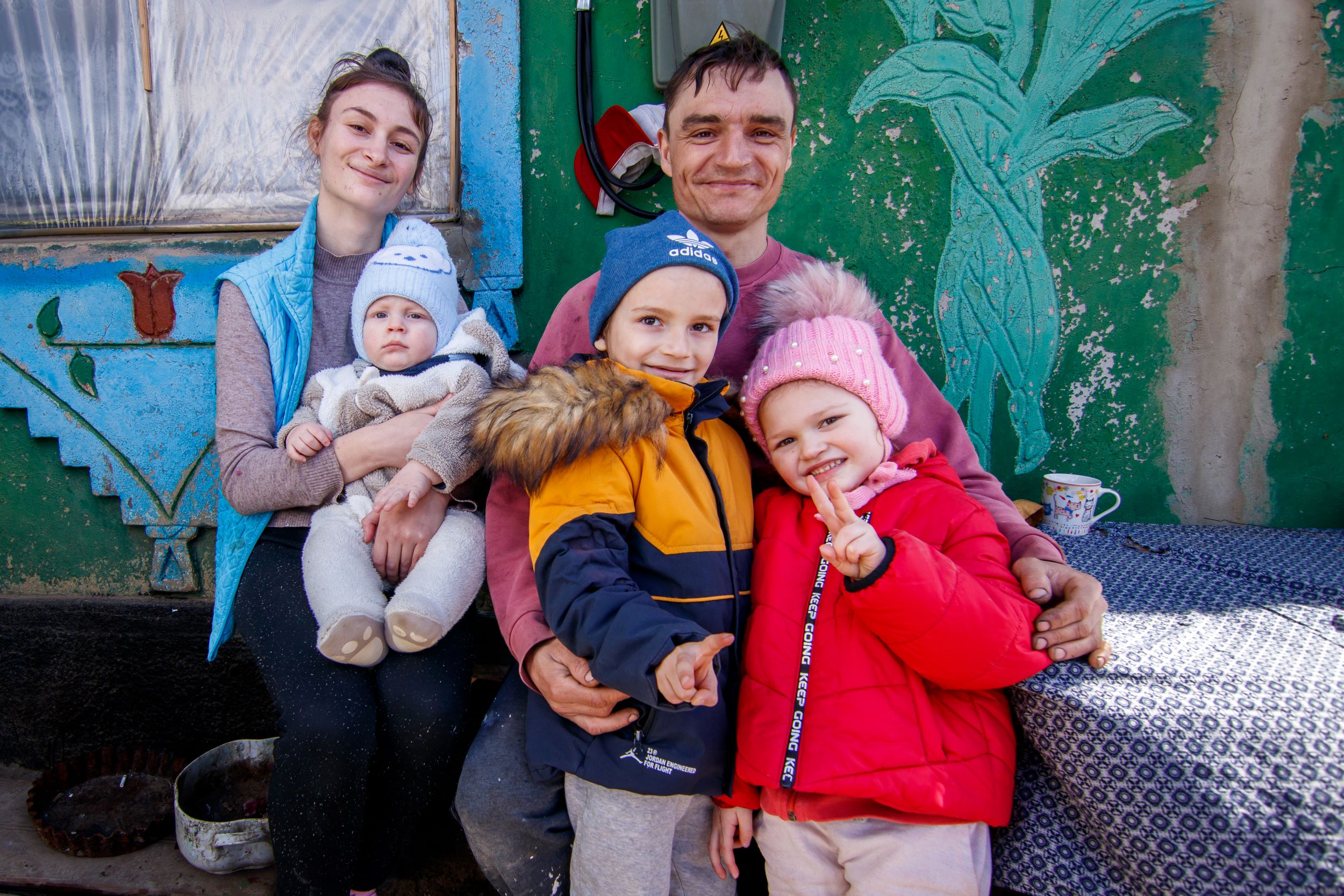  Two parents pose with their three children in front of the green and blue walls of a house