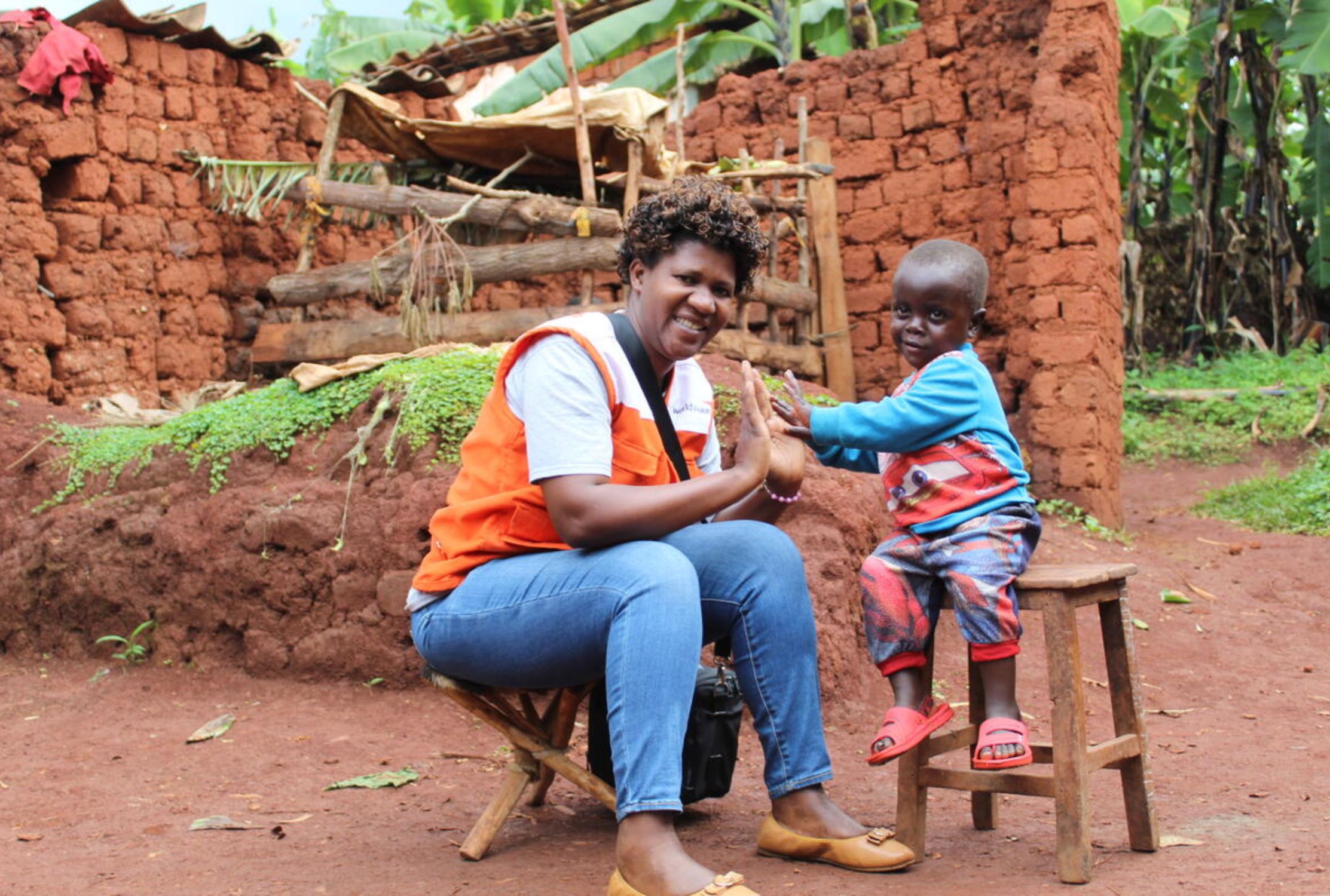 Young boy from Burundi sits on a stool, high-fiveing a local World Vision staff member