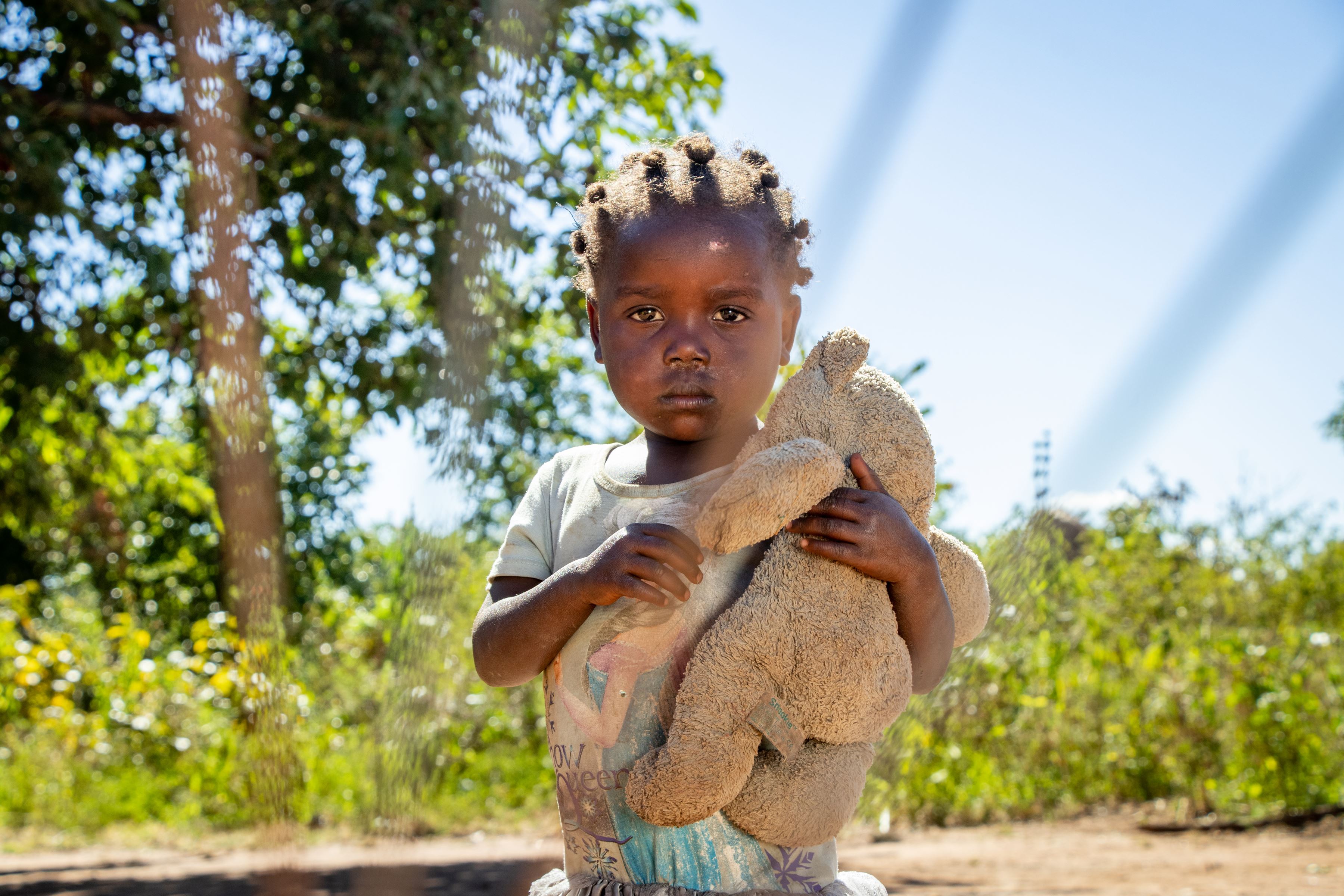 A little girl stands in front of grass and a tree clutching a teddy bear. 